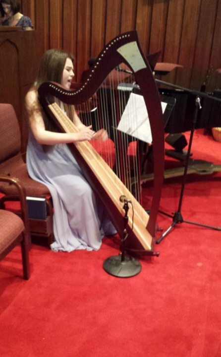 Young girl in a light dress sitting on a chair, playing a large harp in a room with wooden walls and red carpet.