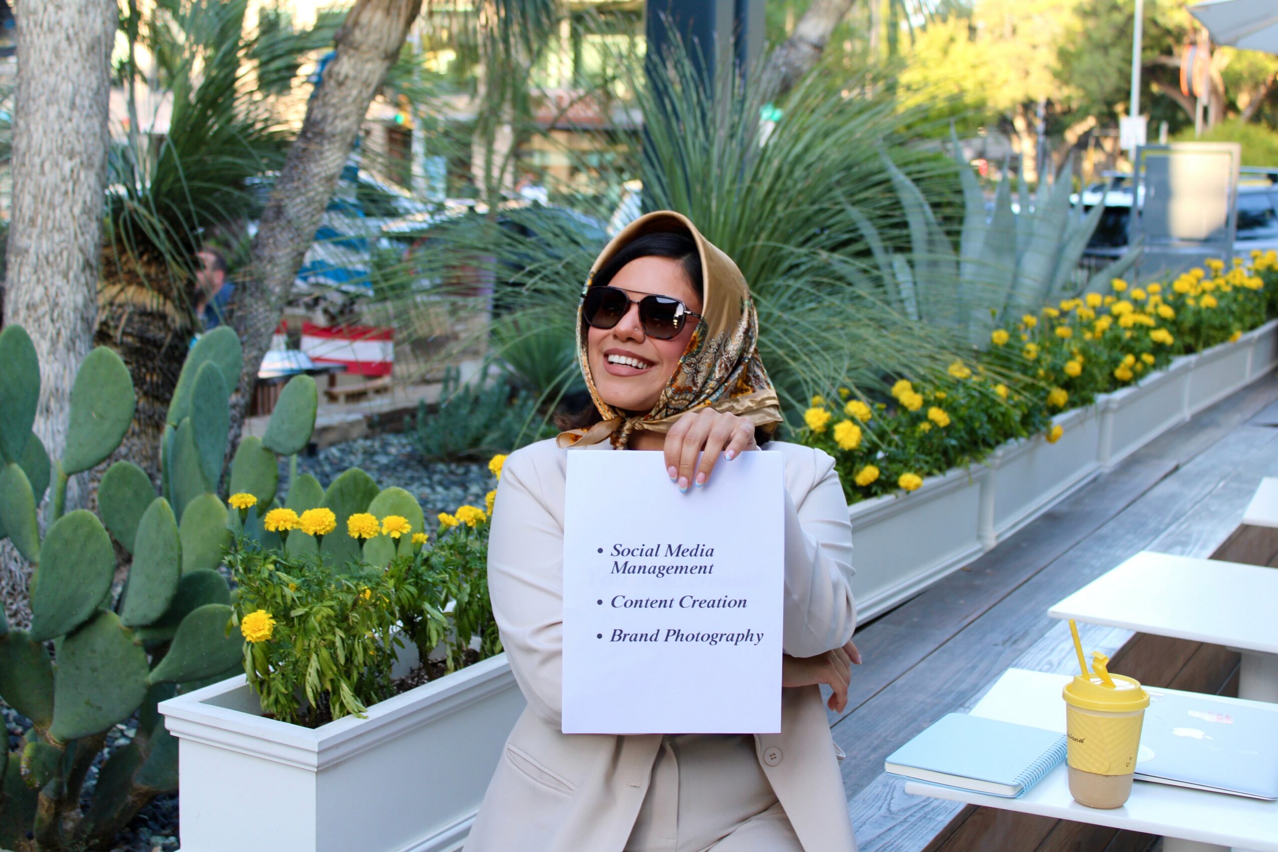 Woman smiling outdoors holding a sign, wearing sunglasses and a headscarf, with plants and flowers in the background.