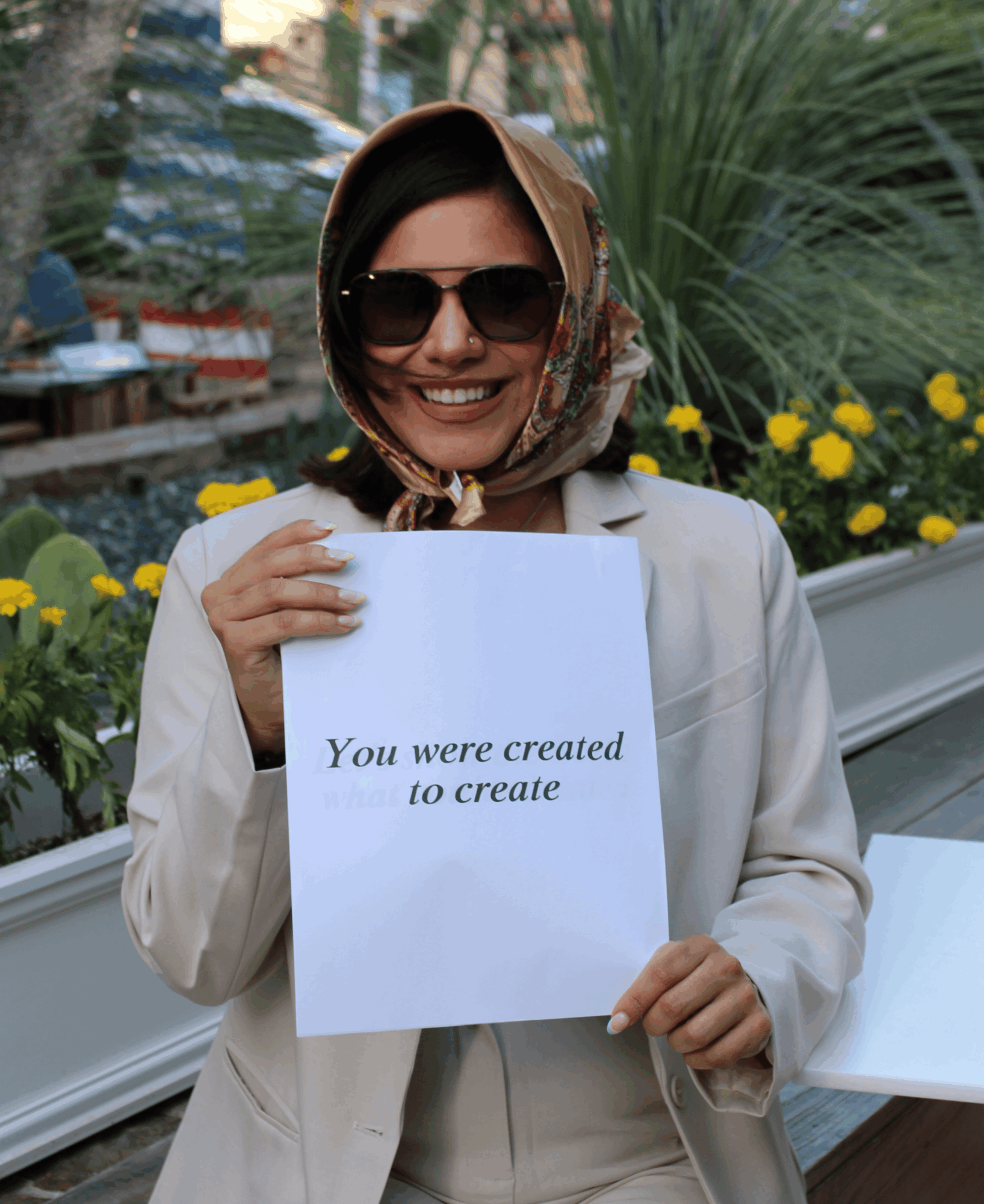 Smiling woman wearing sunglasses and a headscarf holding a sign with text, outdoors with plants and flowers in background.