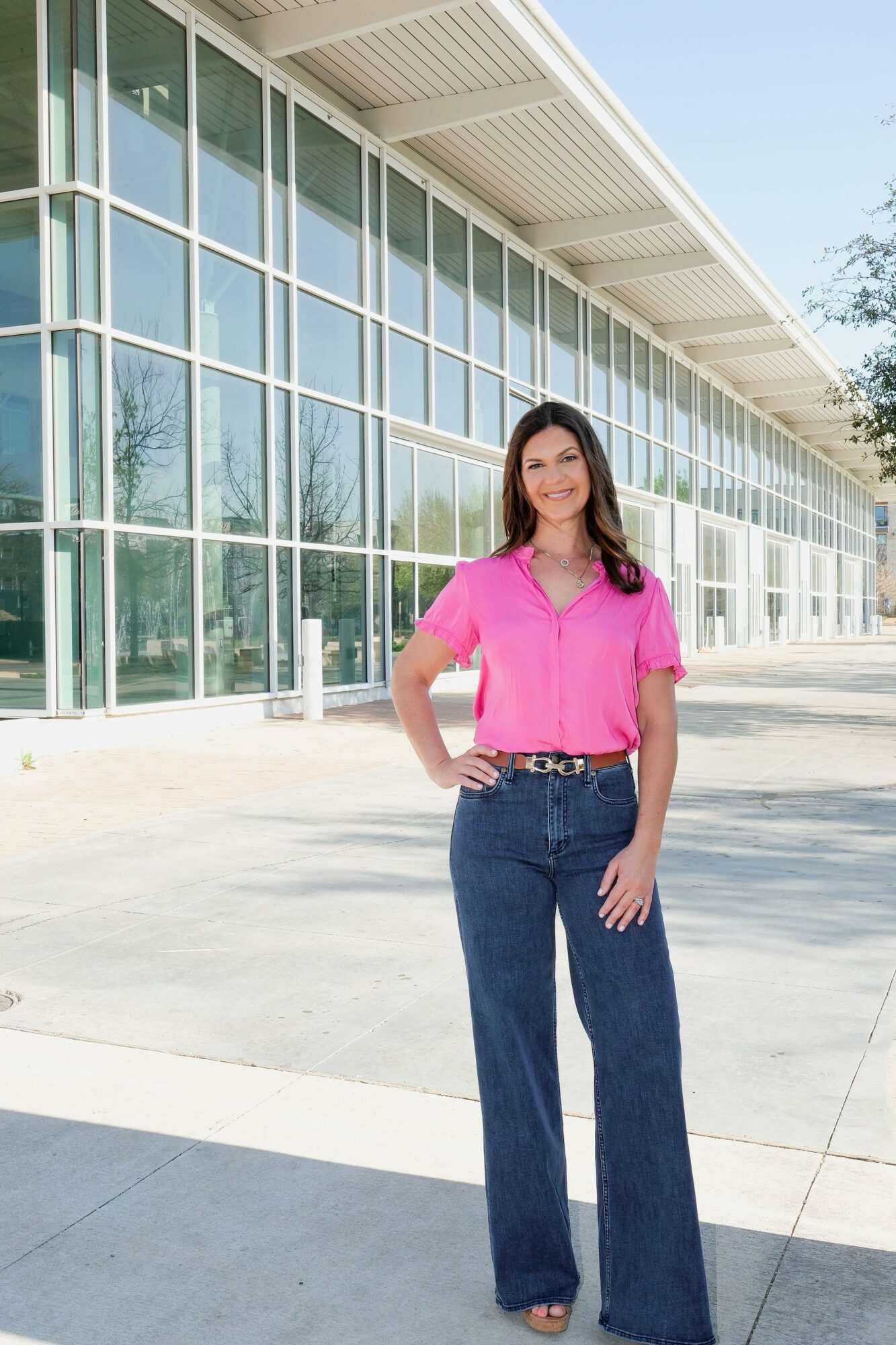 Woman in pink top and jeans standing outside modern glass building, sunny day.