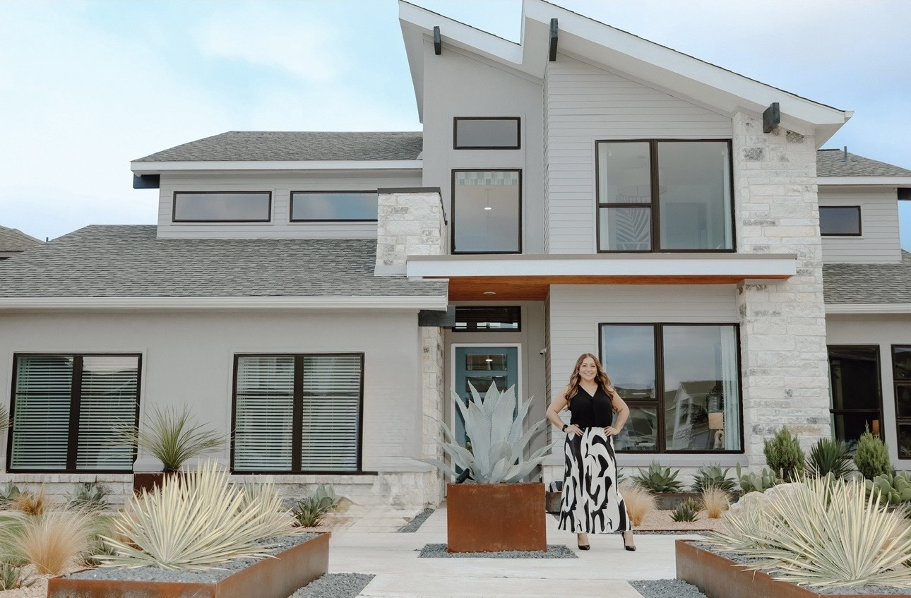 Woman standing outside modern house with large windows and desert landscaping, blue sky background.
