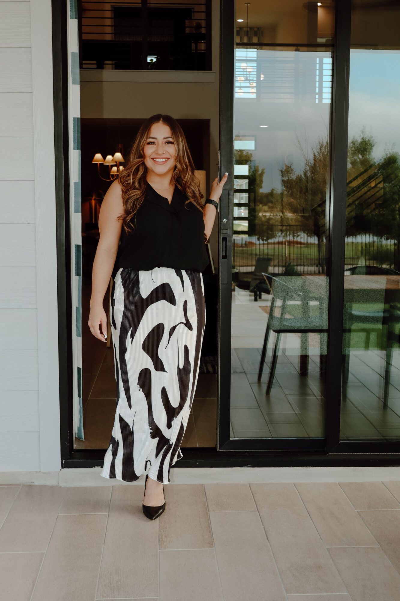 Woman standing in front of glass door, smiling, wearing a black top and patterned pants, indoors and outdoors visible.