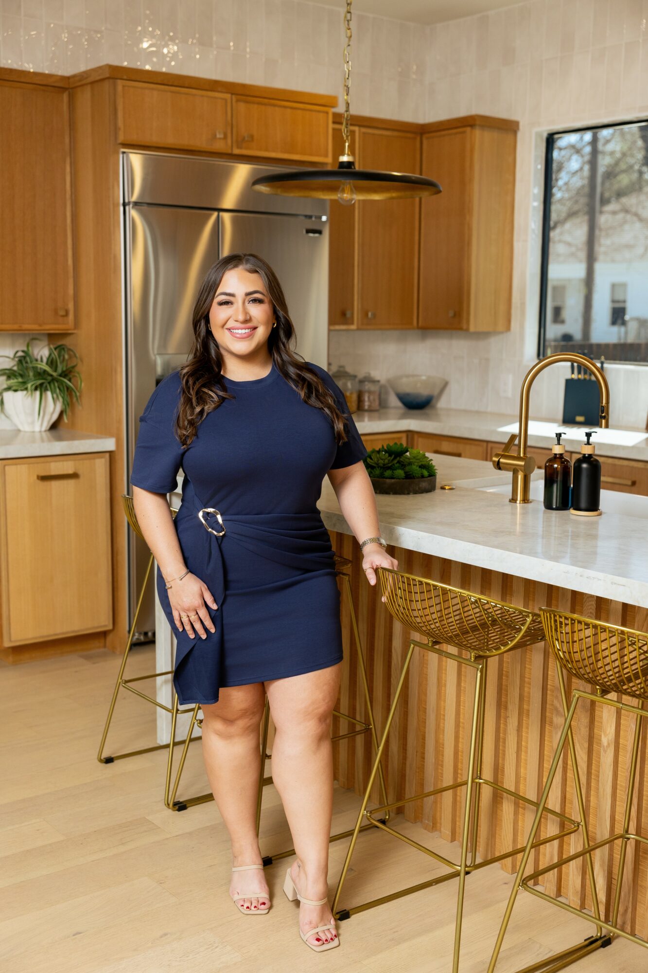 Woman in navy dress standing in a kitchen with wooden cabinets, marble island, and gold bar stools, smiling at the camera.