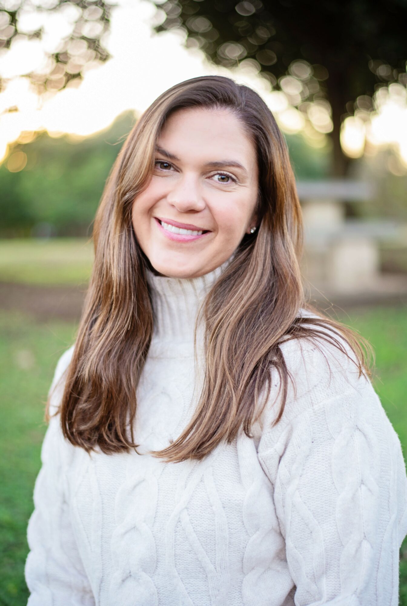 Young woman with long brown hair smiling outdoors in a park, wearing a white sweater, with trees and sunlight in background.