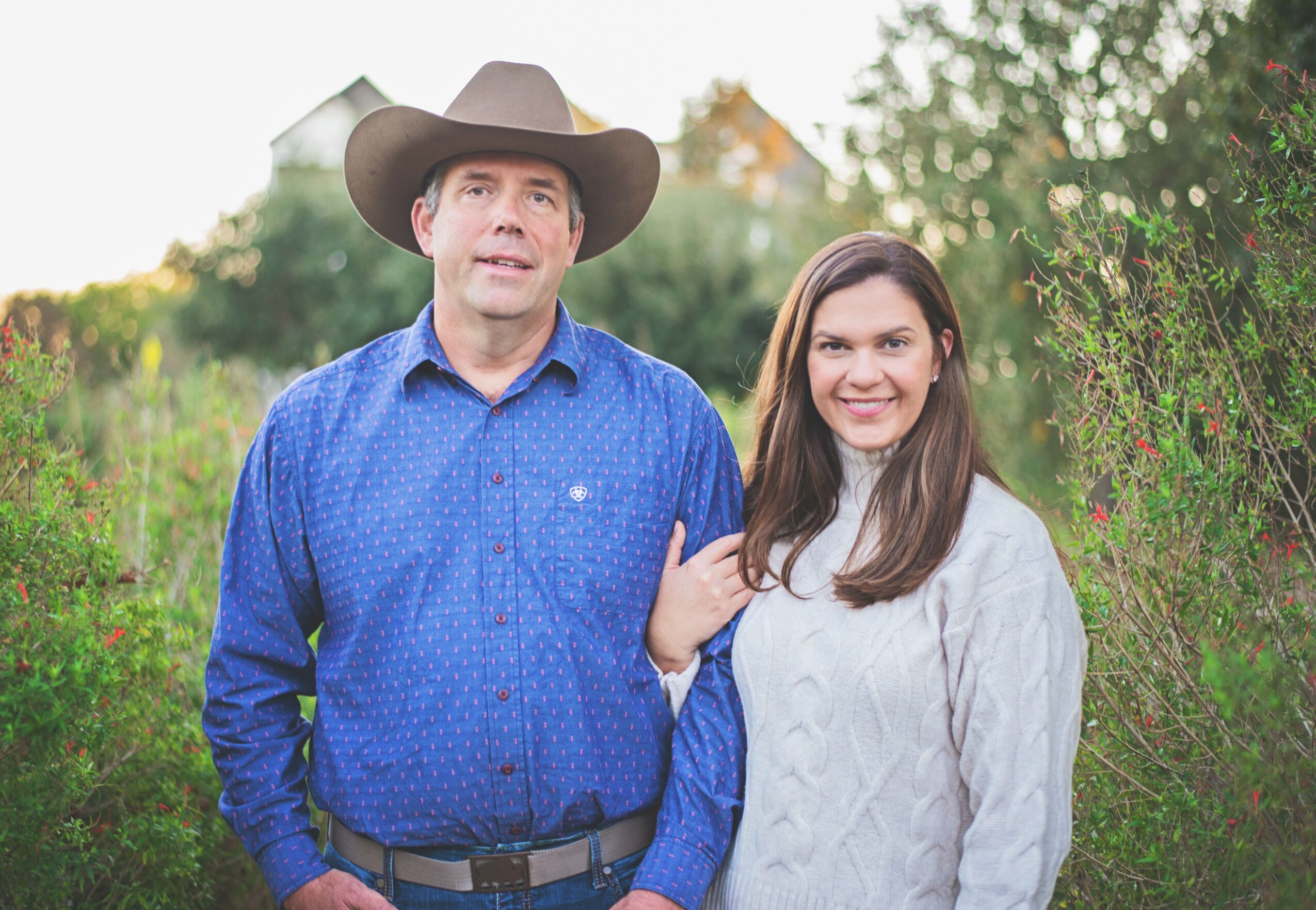 A man wearing a cowboy hat and a woman standing outdoors with greenery, smiling, and holding hands.