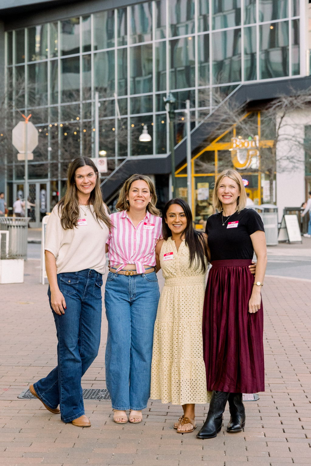 Four women standing outdoors in front of a modern glass building, smiling, dressed casually, with trees and streetlights in background.