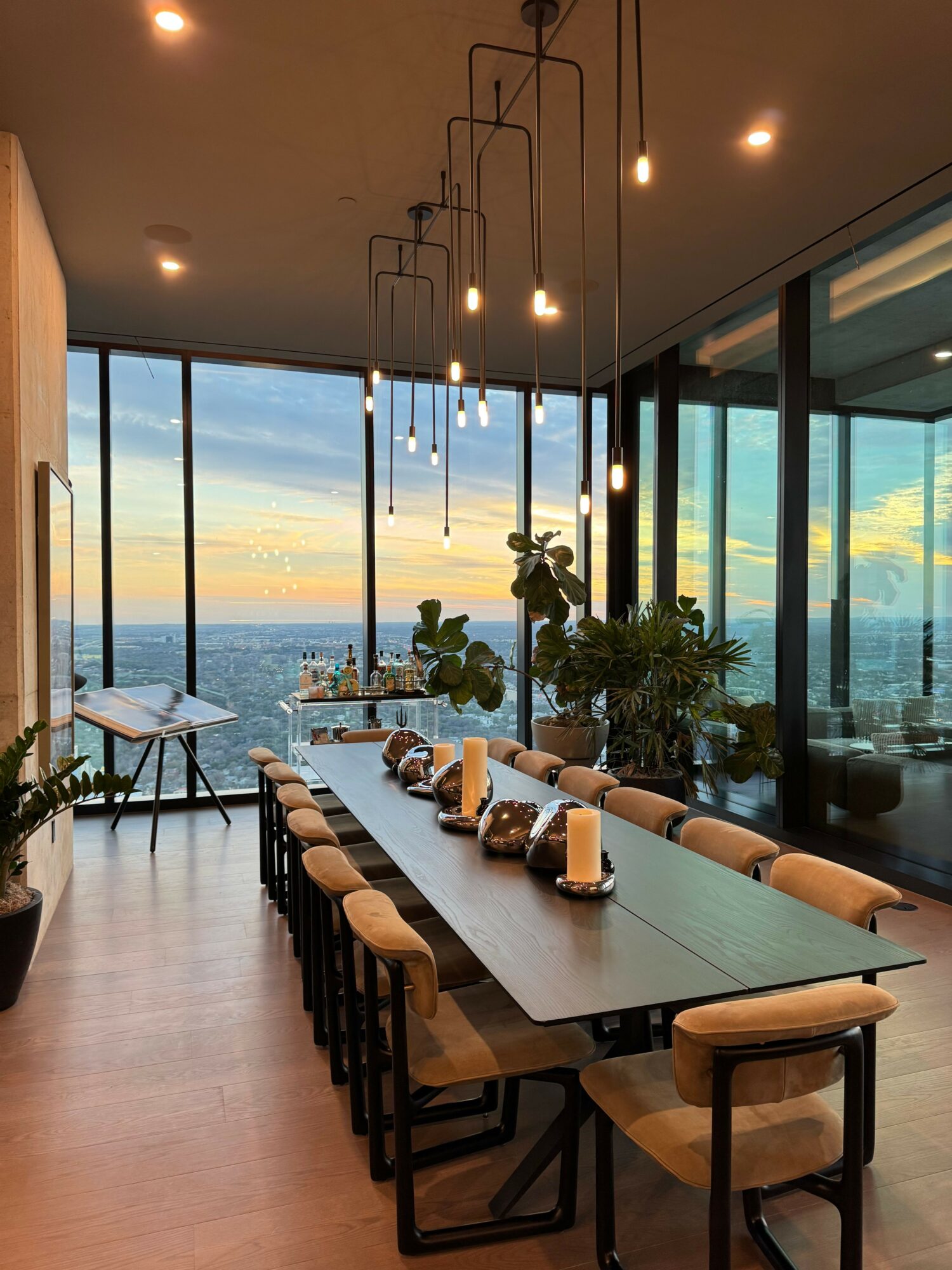 Modern dining area with a long table, chairs, and large windows showing a city view, plants on the table.