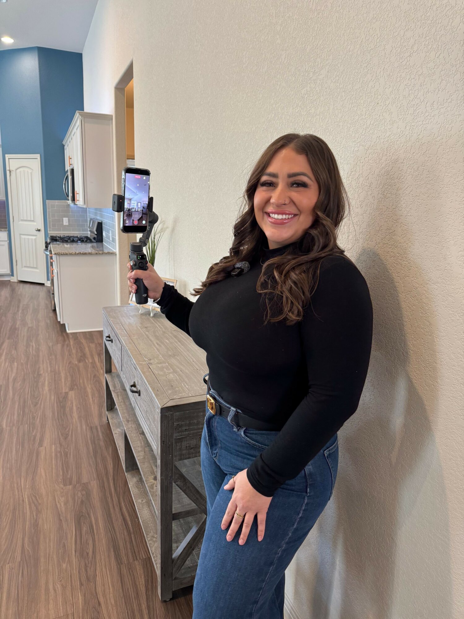 Woman with long brown hair smiling, holding a phone, standing next to a table with a laptop, in a room with beige walls.