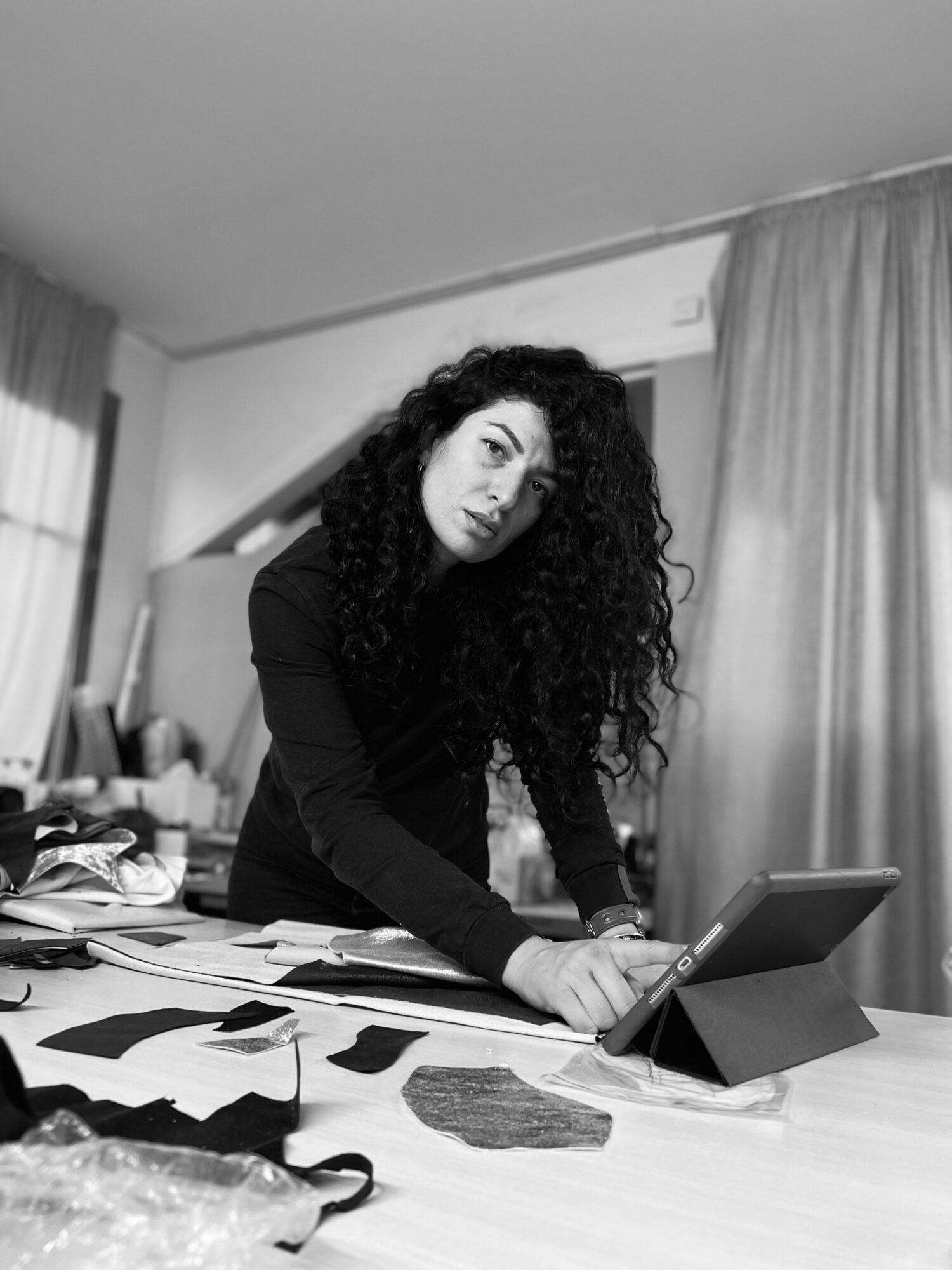 Woman with curly hair working on a tablet at a cluttered desk in a room with curtains.