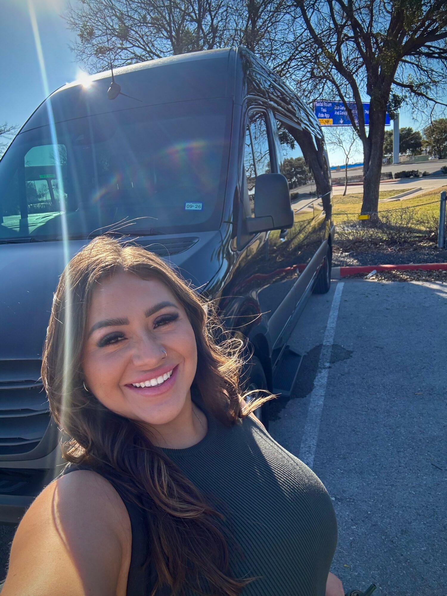 Smiling woman with long hair in a parking lot, sunny day, trees, and a van behind her.