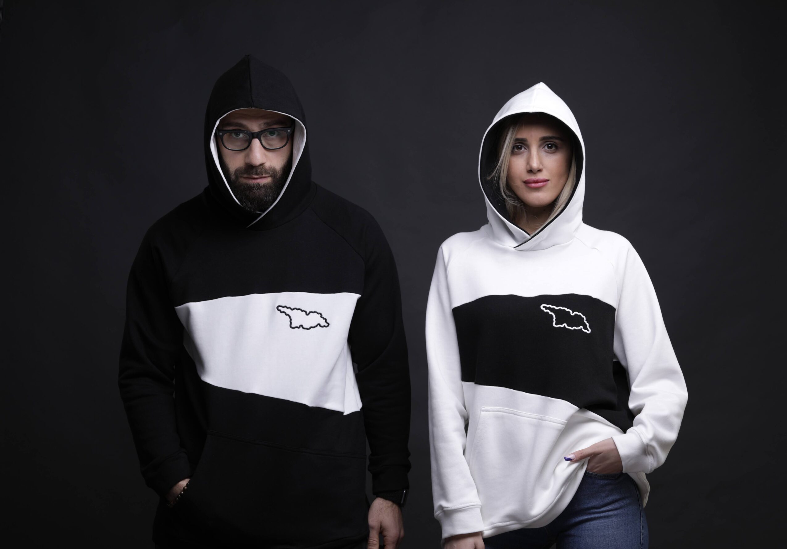 Two people wear hoodies with a black and white color block design and a logo, standing against a dark background.