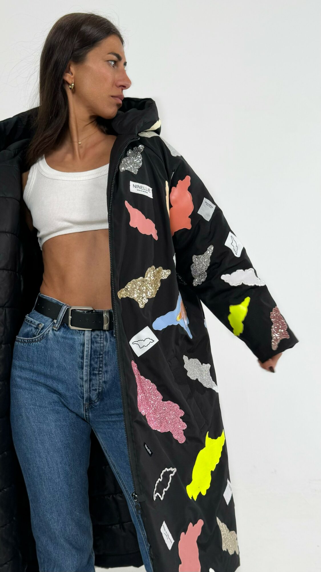 Woman with long dark hair wearing a white crop top, blue jeans, and a black coat with colorful abstract patterns, standing sideways against a plain background.