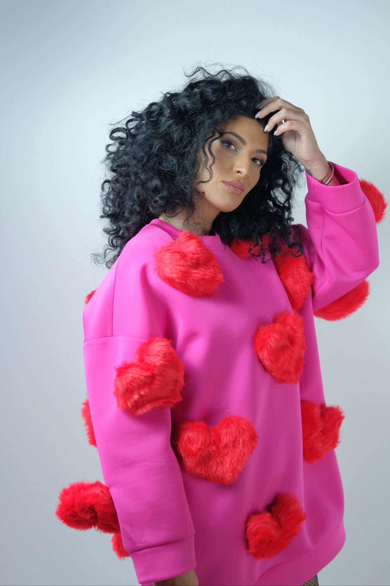 Woman with curly hair wearing a pink sweater with red pom-poms, touching her hair, against a plain background.