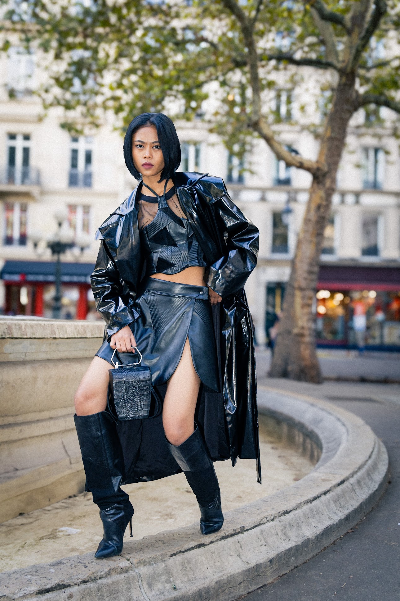 Woman in black leather outfit standing on a city street with trees and buildings in background.