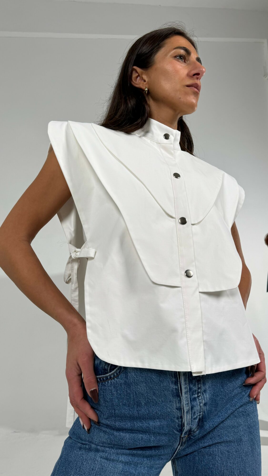 Woman in white sleeveless top with ruffled shoulders and blue jeans, standing sideways with hands in pockets.