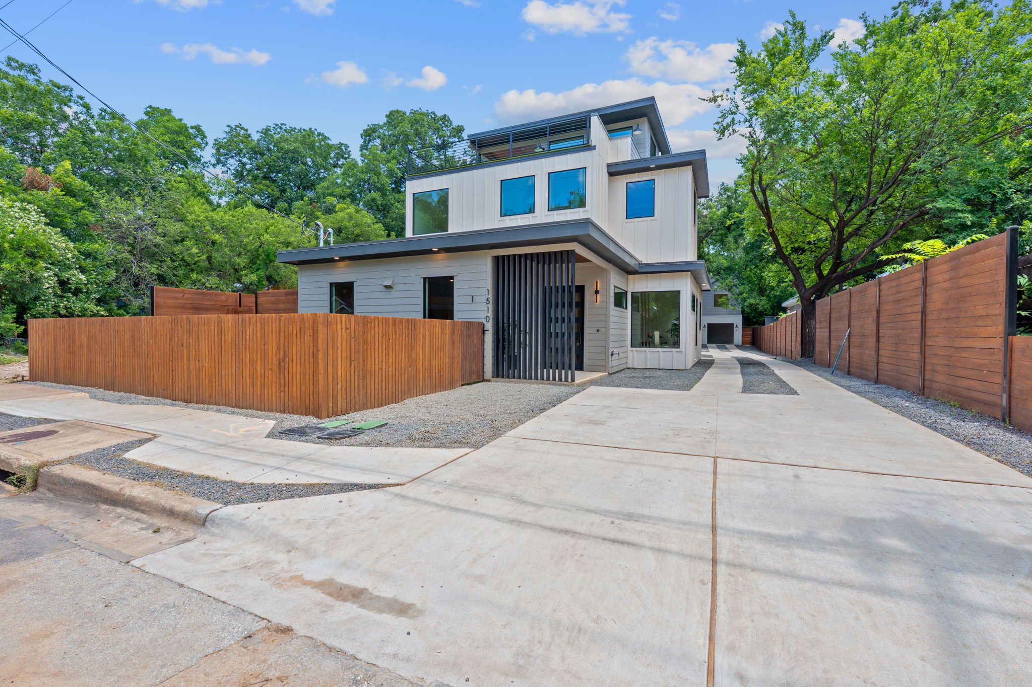 Modern multi-story house with large windows, surrounded by trees and a wooden fence, with a concrete driveway.