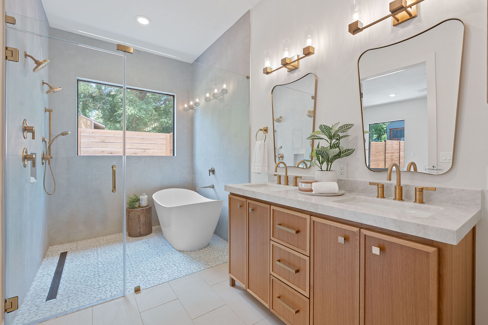 Modern bathroom with a bathtub, large window, double vanity with two mirrors, and a plant, illuminated by wall lights.