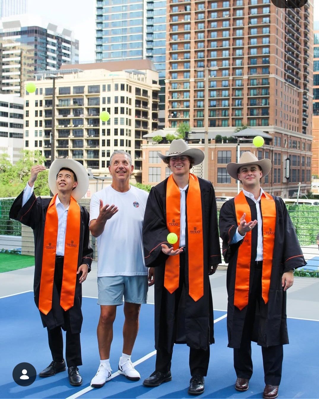 Four people standing outdoors in front of tall buildings, two in graduation gowns and caps, one in casual clothes, all holding tennis balls.