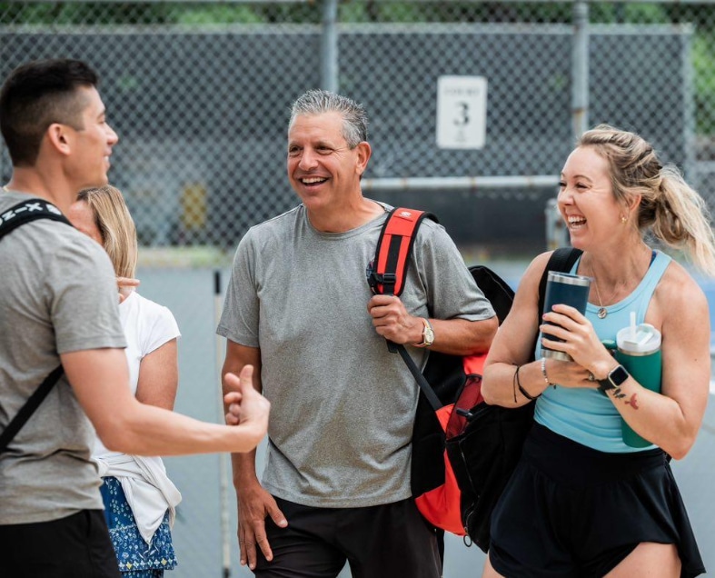 Four people smiling and talking outdoors near a tennis court, two holding drinks, one with a backpack.
