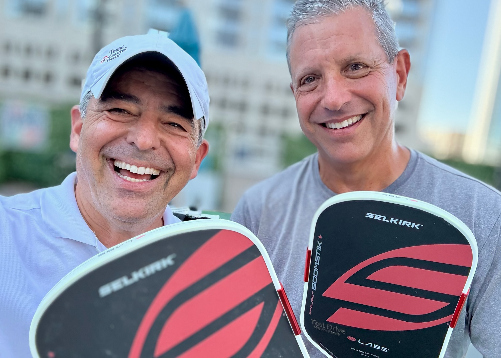 Two smiling men holding pickleball paddles outdoors with buildings in the background.