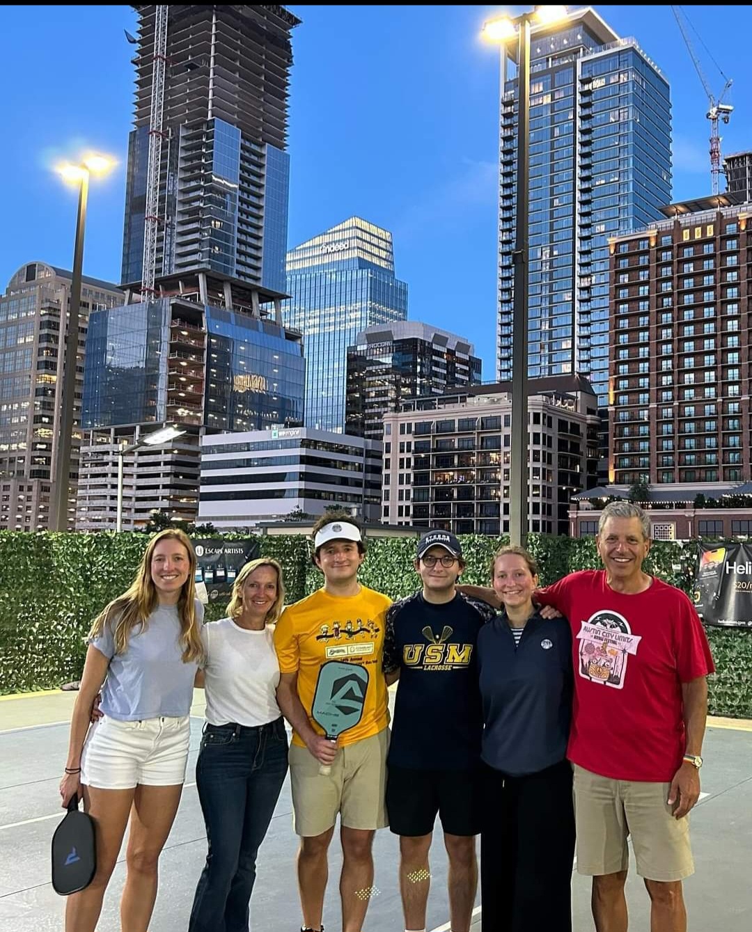 Group of six people standing outdoors in front of city skyscrapers, smiling, with a green hedge behind them.