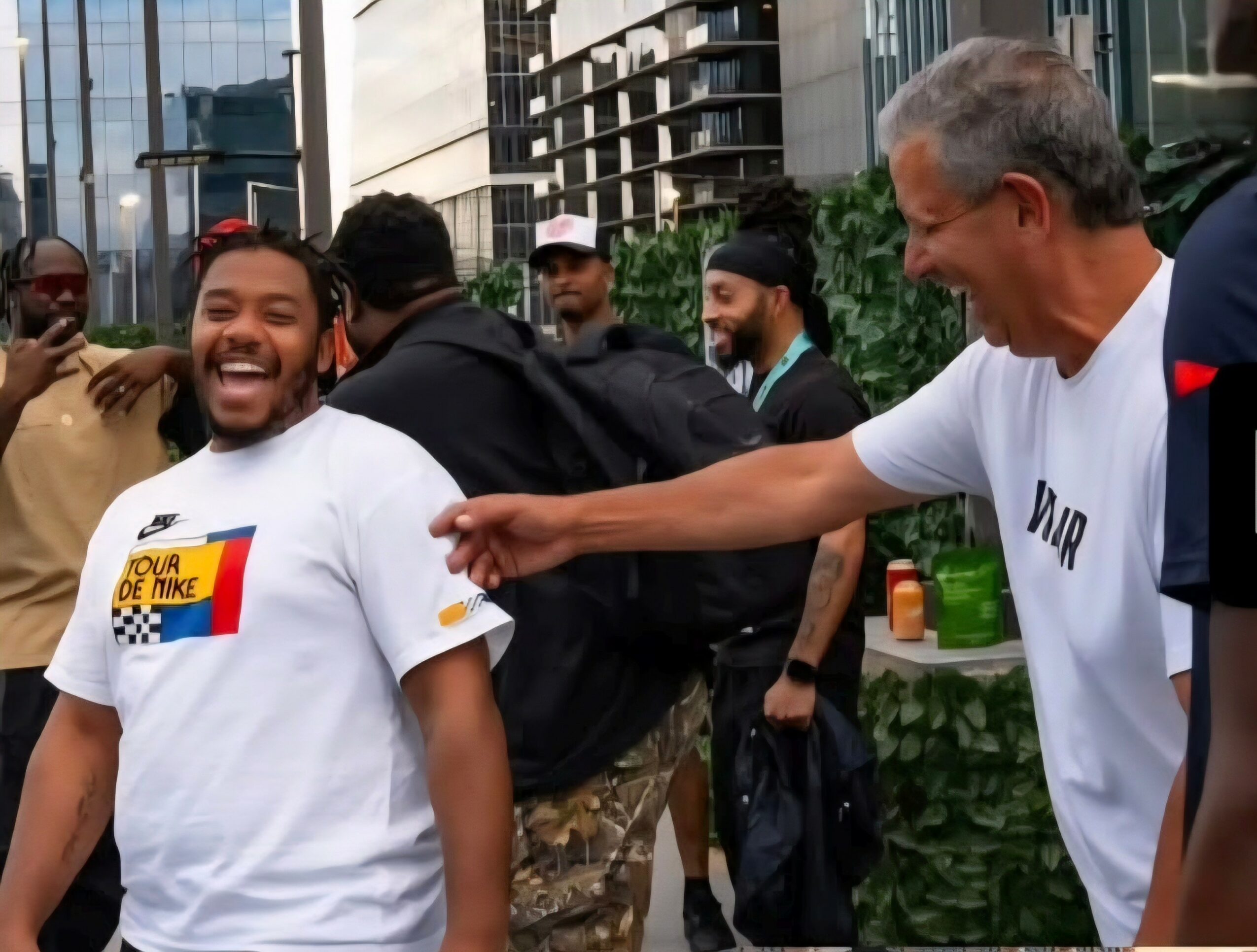 Two men laughing and smiling outdoors in an urban setting with several people around, some holding backpacks, in front of tall buildings.