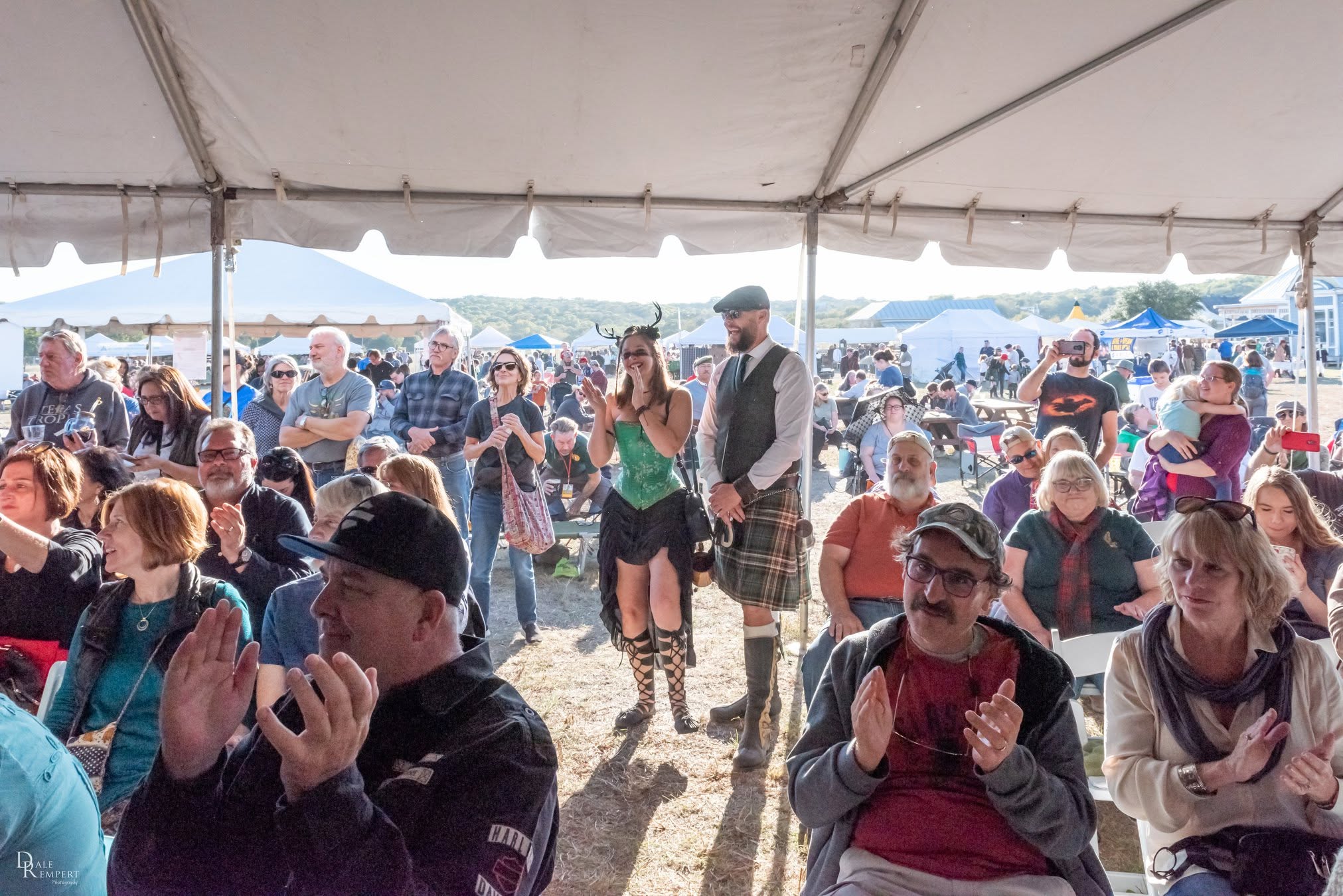Crowd of people gathered under a tent at an outdoor event, some clapping and socializing, with vehicles and tents in the background.