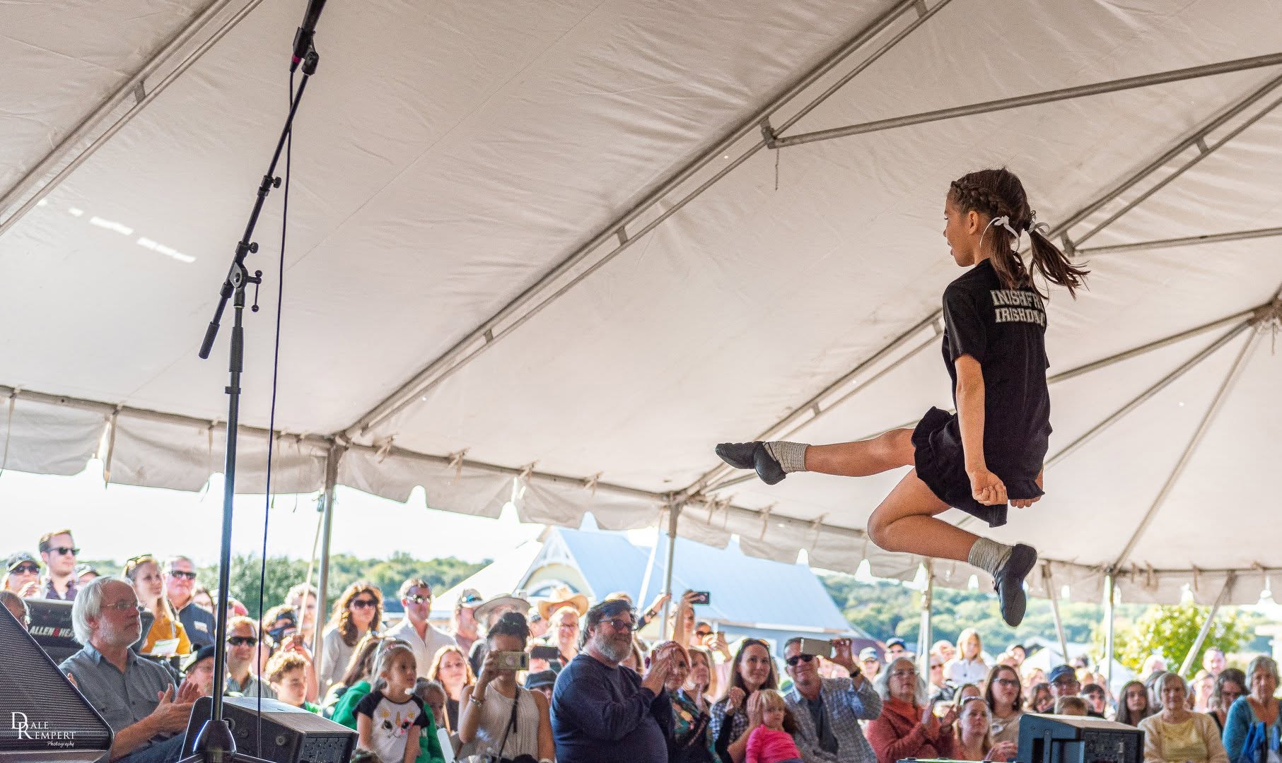A young girl performs a jump on a tightrope under a large tent, with an audience watching below.