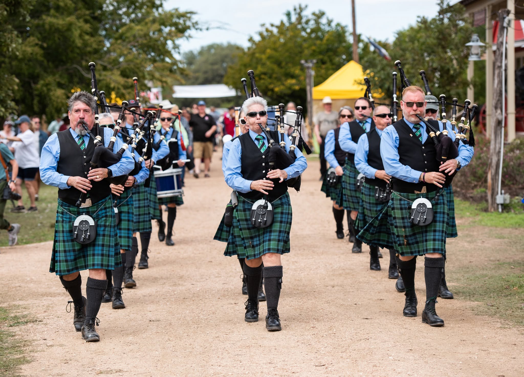 Group of people in traditional Scottish attire playing bagpipes and marching outdoors during daytime.