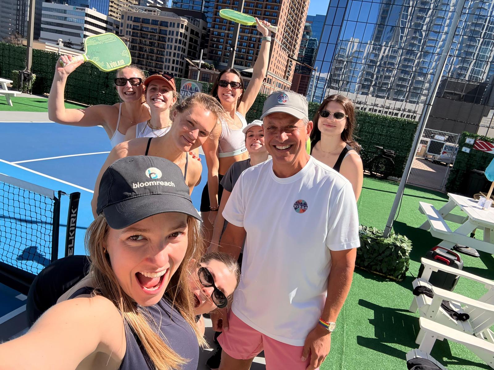 Group of seven people smiling and holding pickleball paddles on a court with city buildings in background.