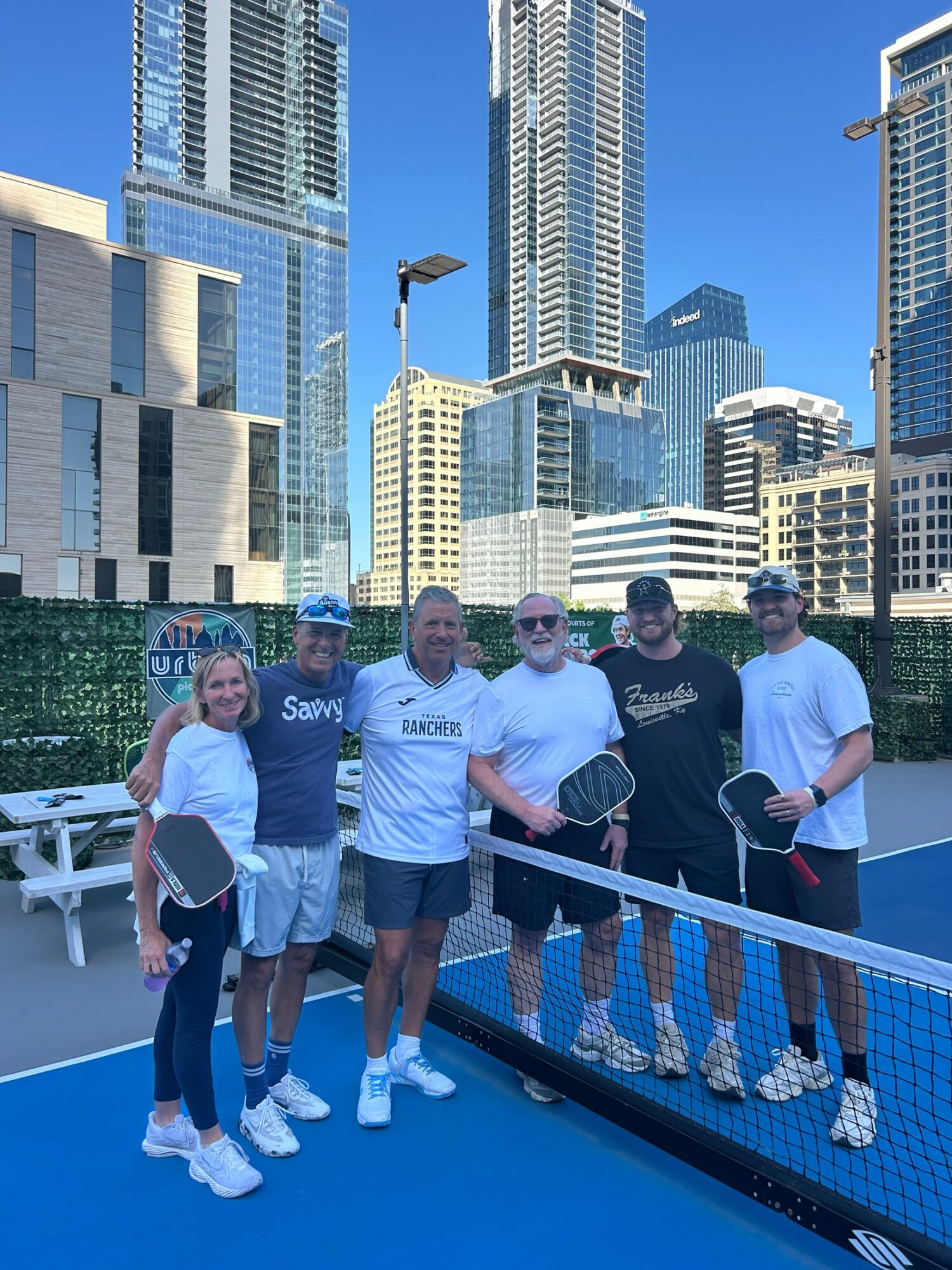 Five people stand on a rooftop tennis court with city skyscrapers in the background, holding tennis rackets, smiling.