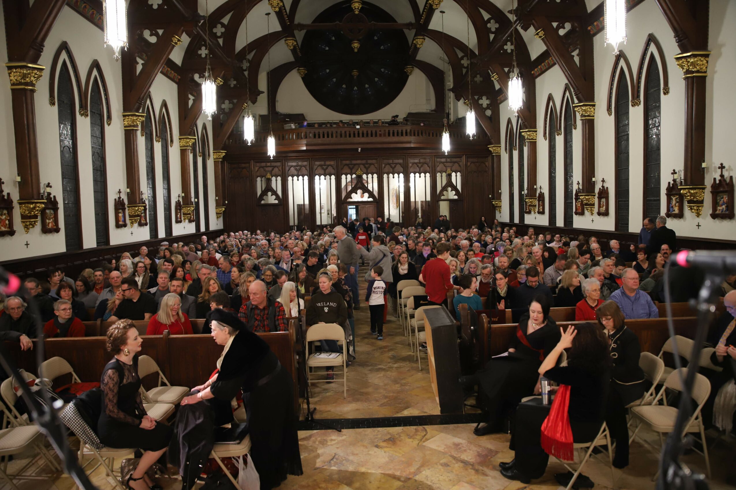 Large hall filled with people seated at tables, high arched windows, ornate ceiling, and chandeliers, with a stage at the front.