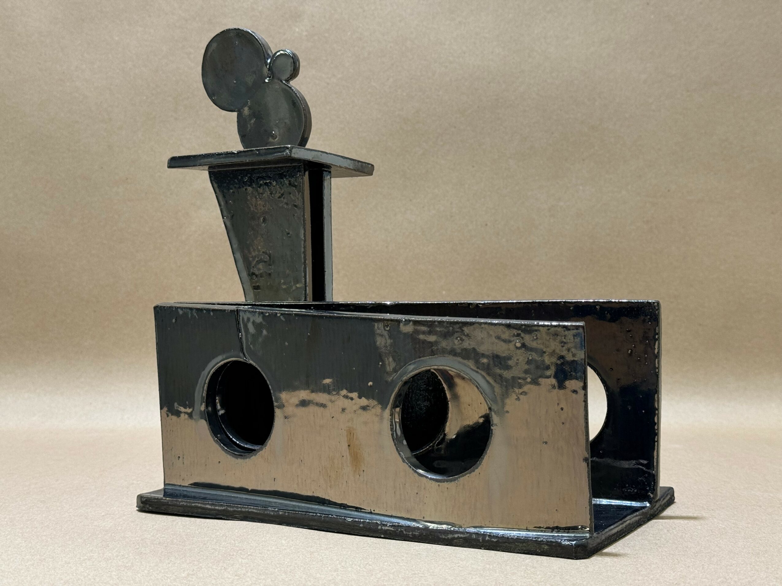 Black metal object with circular holes and a Mickey Mouse-shaped figure on top, placed on a flat surface.