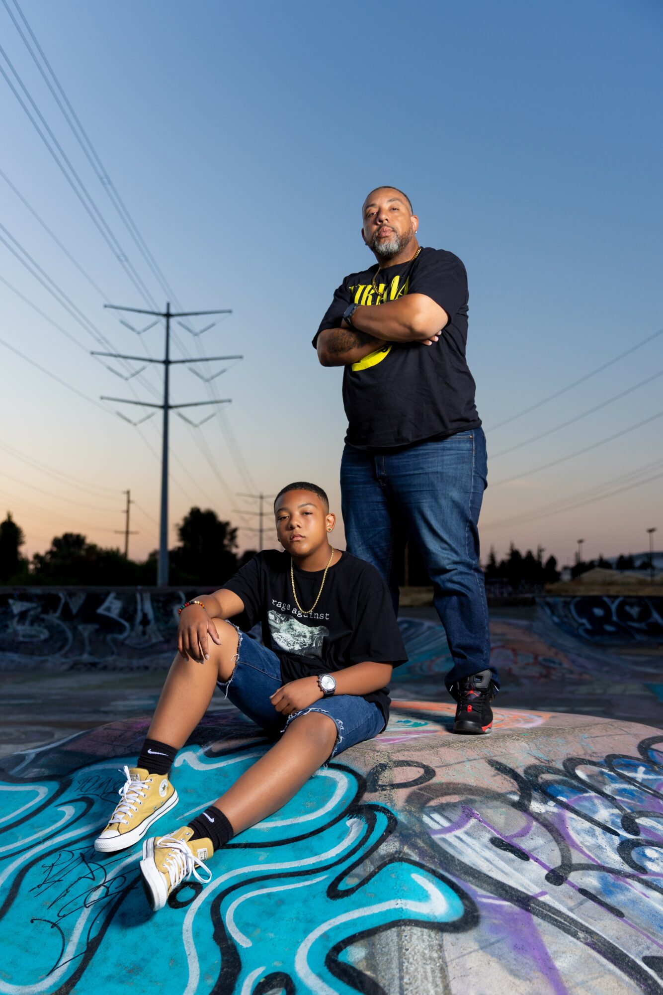 Two men pose on a graffiti-covered surface with power lines and a sunset sky in the background.