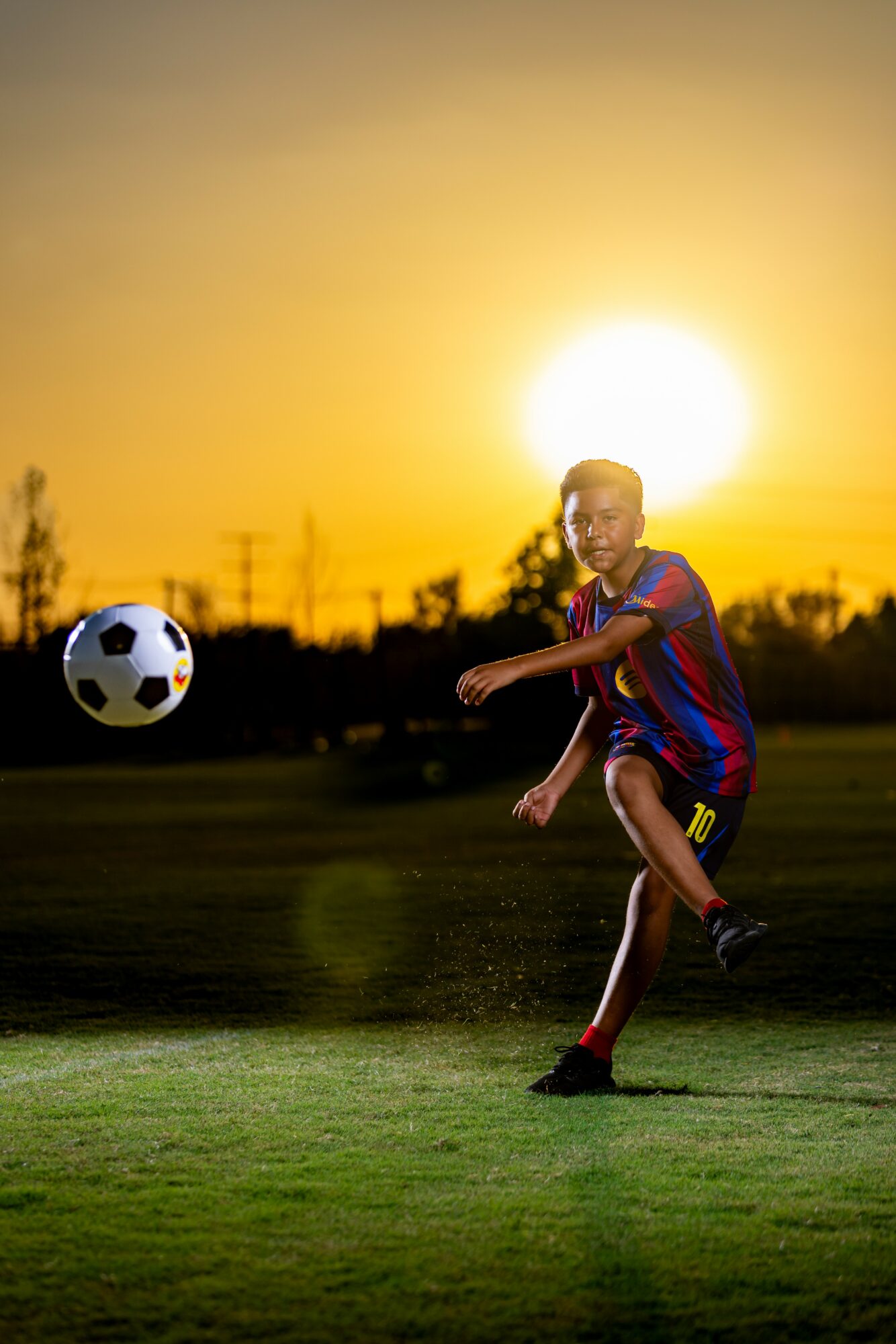 Child playing soccer outdoors during sunset, kicking a ball with a bright sun in the background.