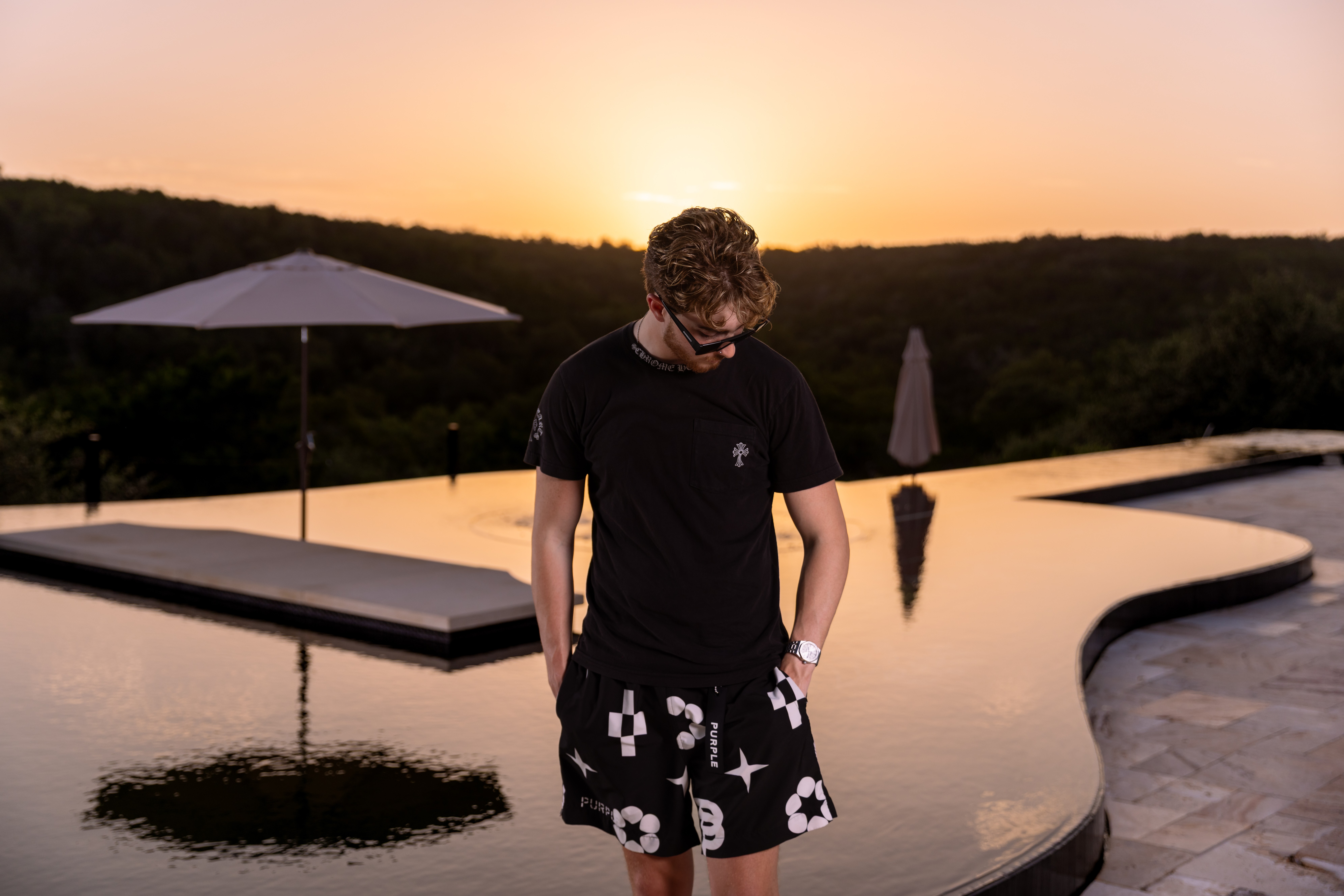 Person standing near a pool at sunset, wearing a black shirt and patterned shorts, with umbrellas in the background.