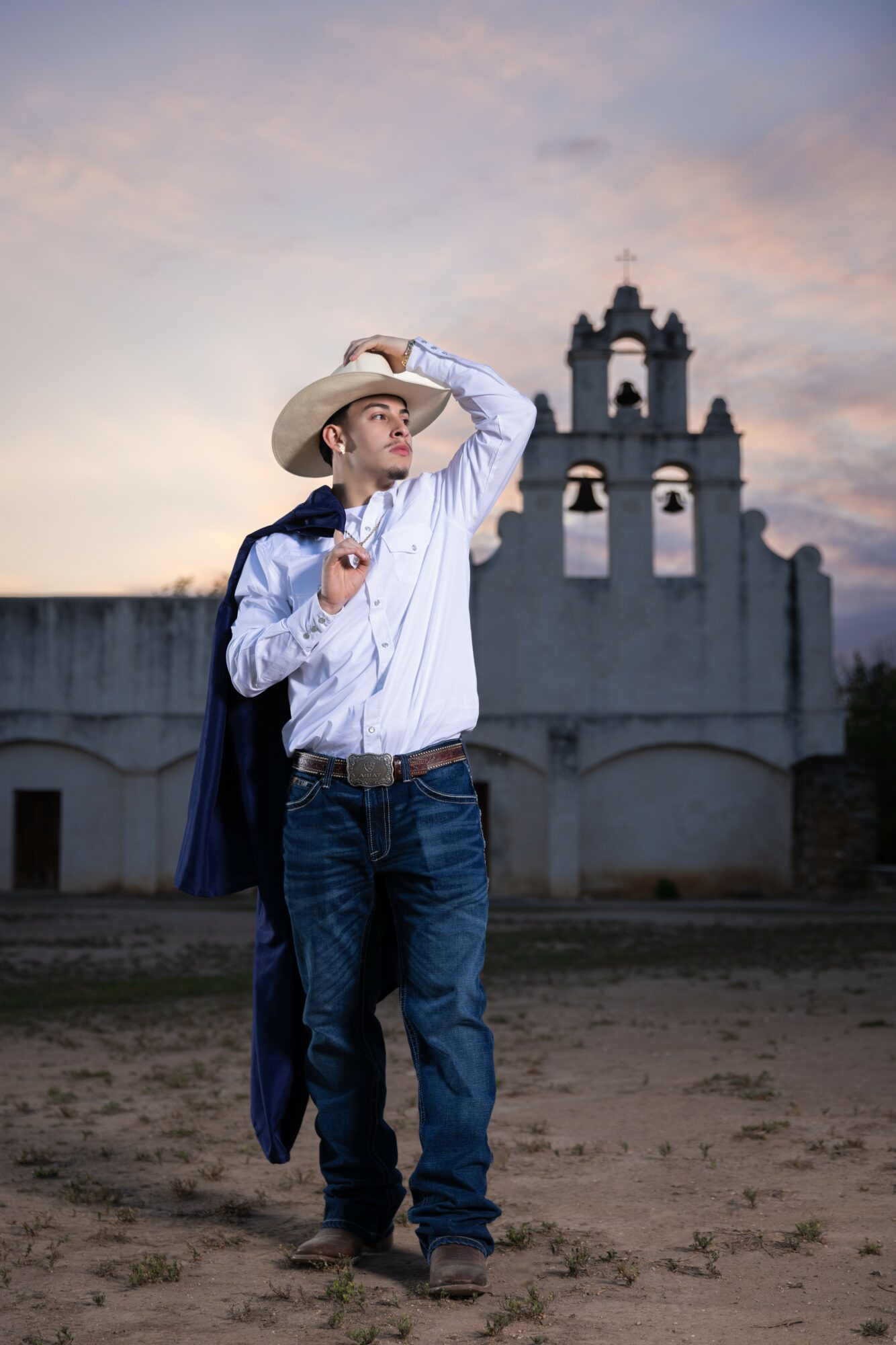 Young man in cowboy hat and jeans stands outdoors near a white church with bell towers at sunset, holding a jacket over his shoulder.