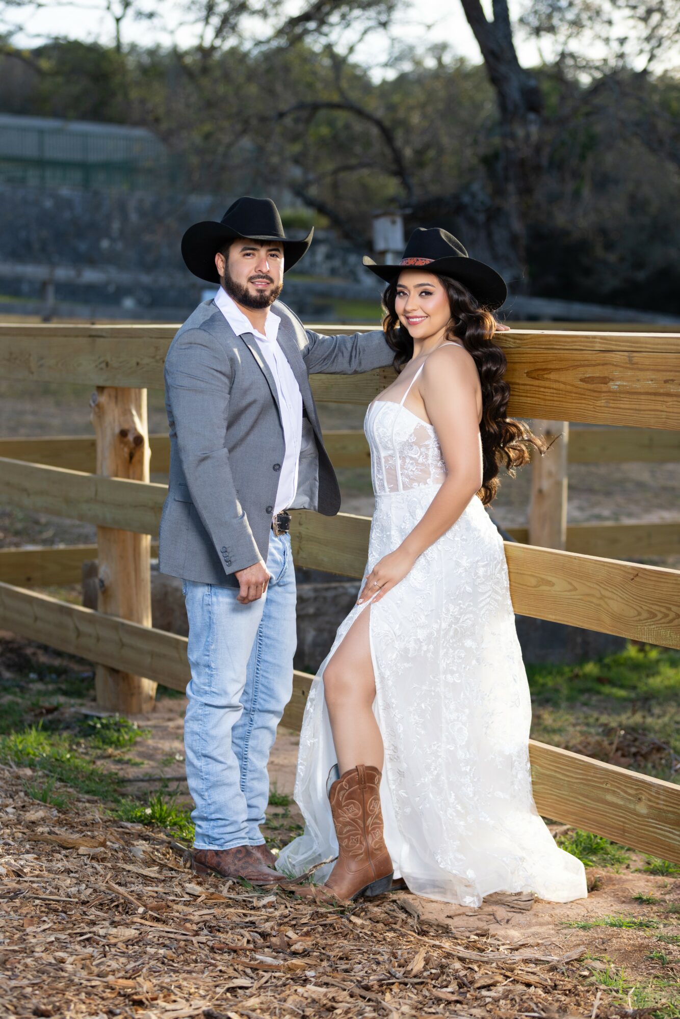 A man and woman stand outdoors near a wooden fence, dressed in stylish clothing, with trees and a fence in the background.
