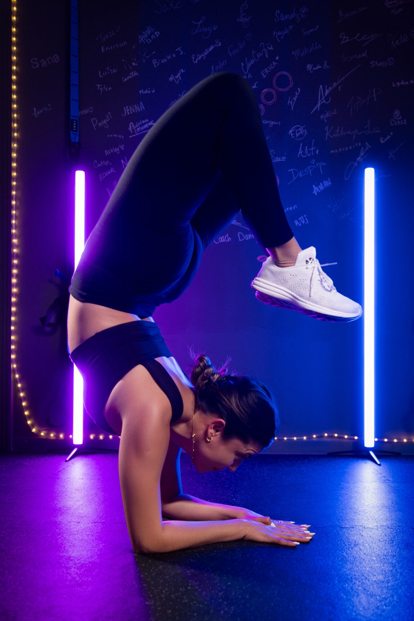 Person performing a headstand with legs bent, in a neon-lit room, wearing black clothing and white shoes.