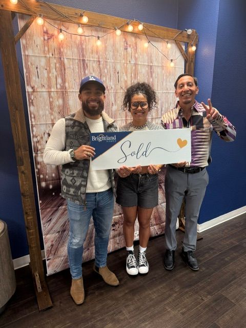Three people standing indoors, holding a sign that says 'Sale' with a heart, in front of a decorated backdrop.