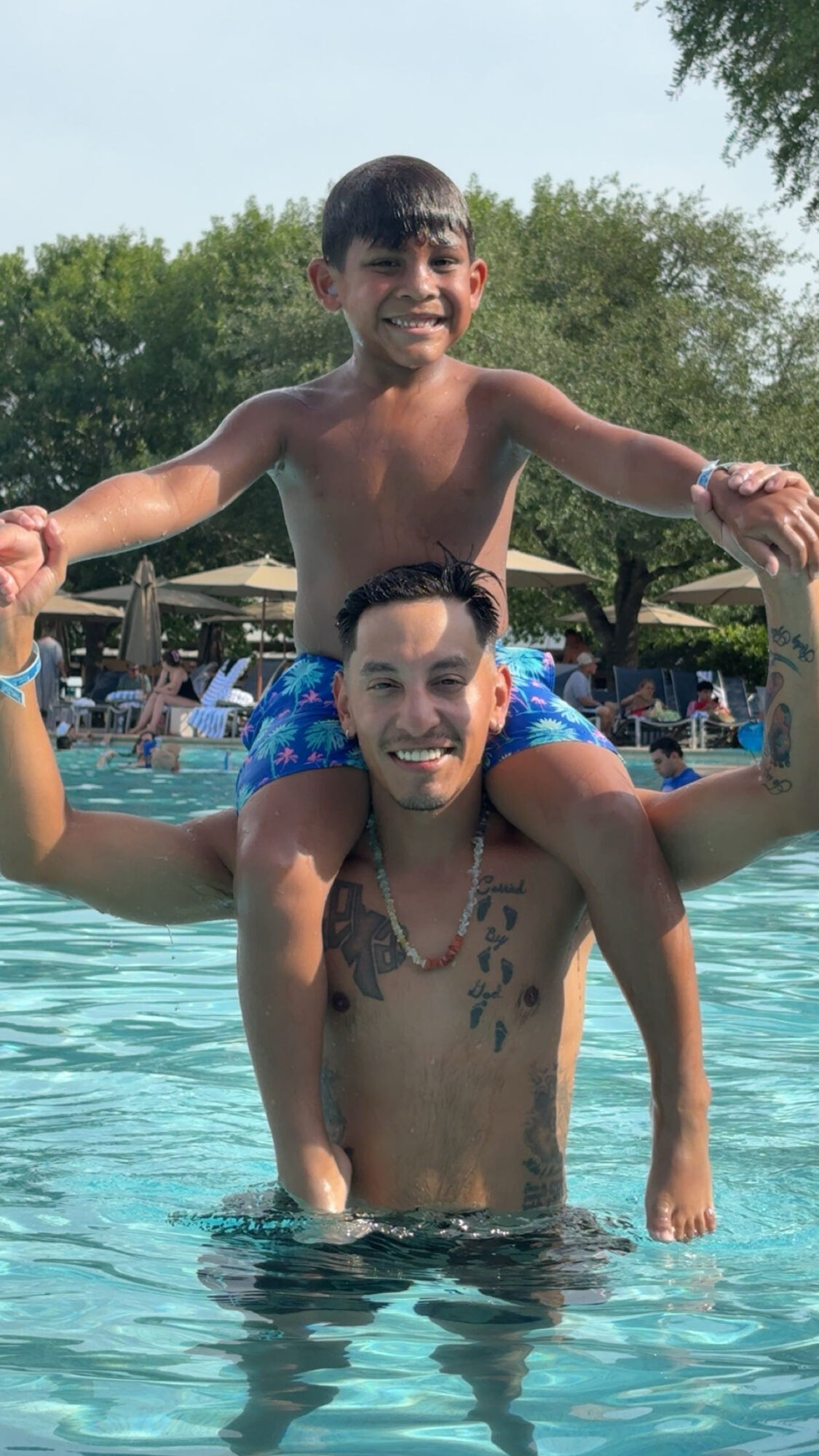 Two boys in a swimming pool, one sitting on the other's shoulders, smiling, with trees in the background.