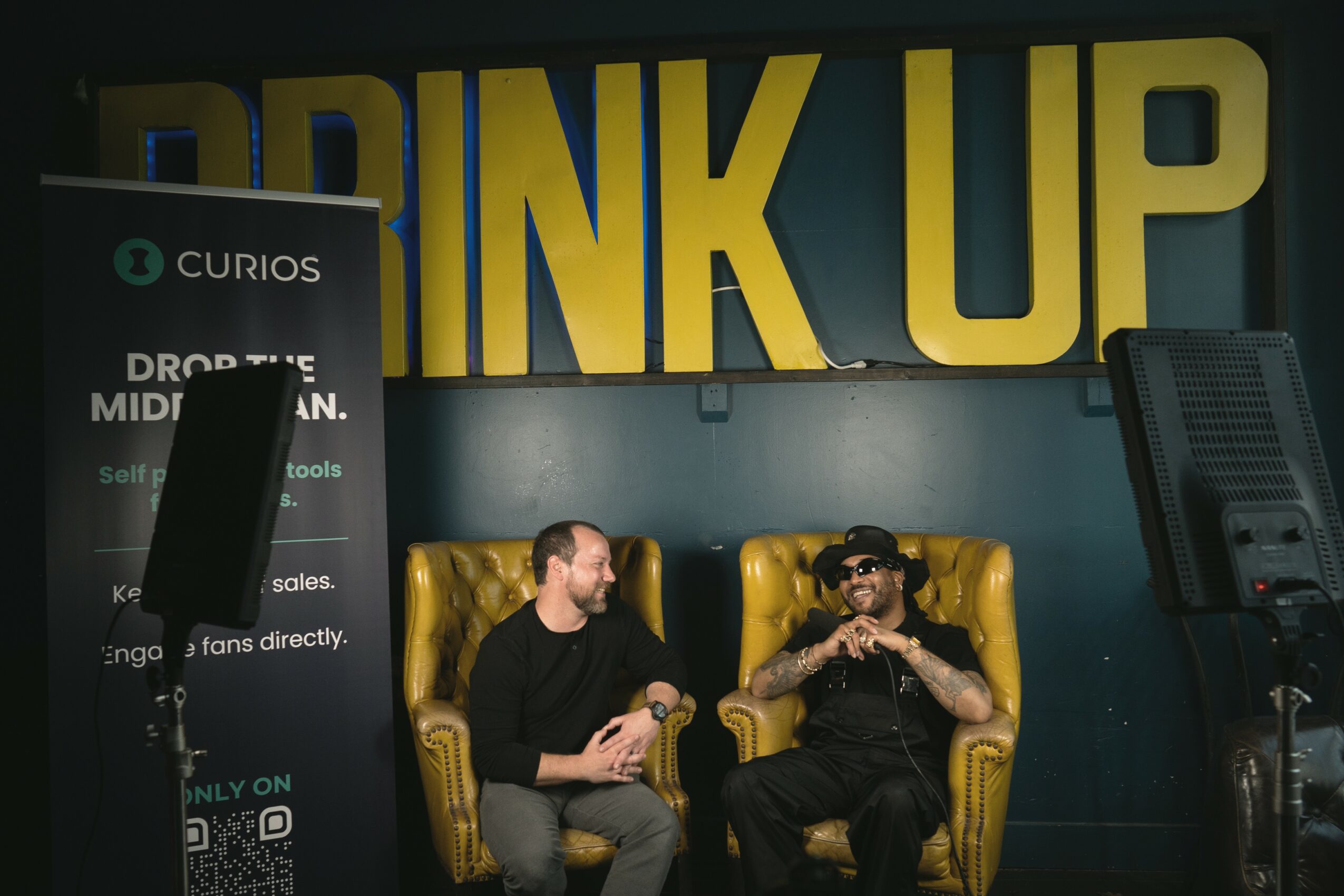 Two men sit on yellow chairs in front of a large yellow and blue sign that says 'DRINK UP'.