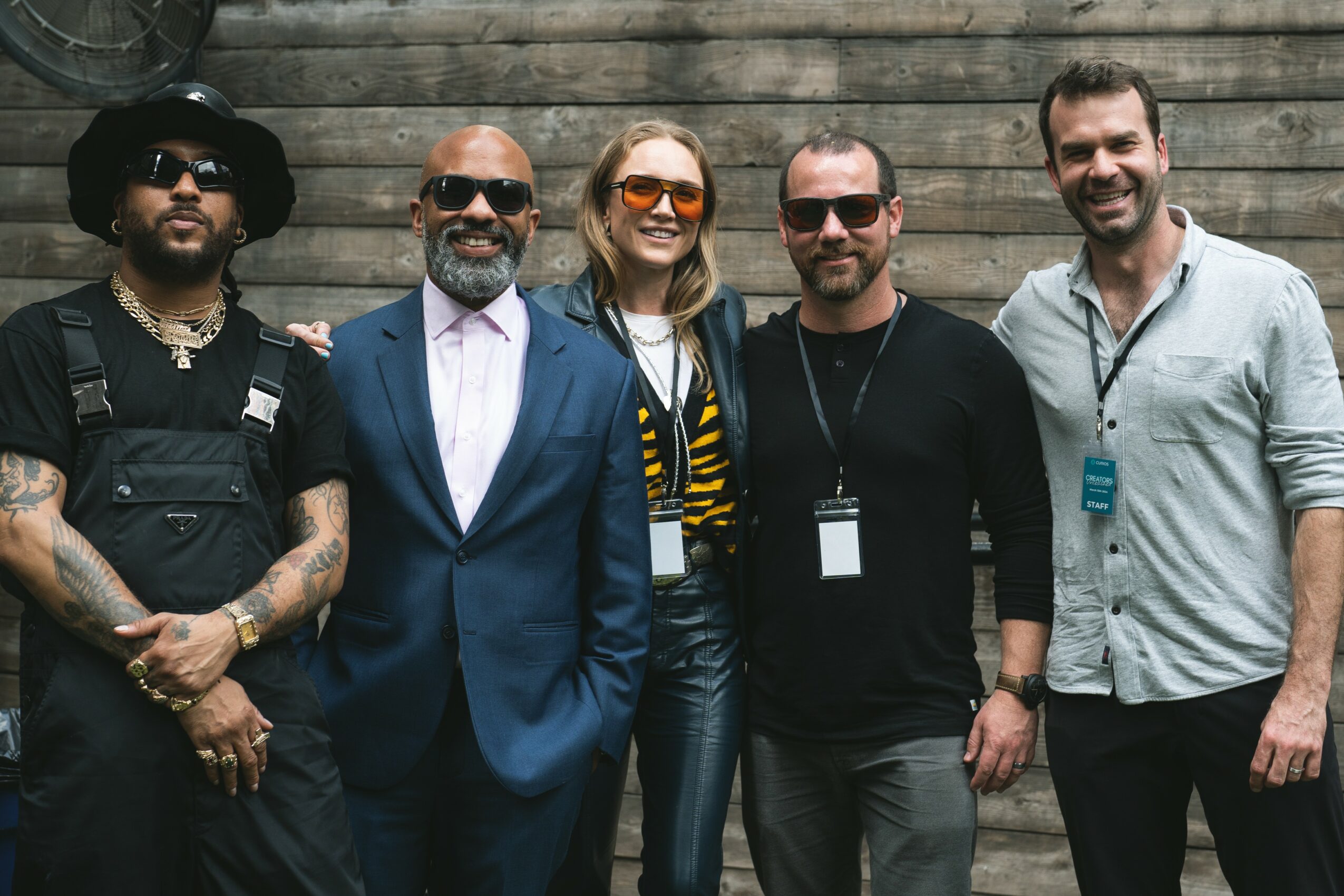 Five people standing in front of a wooden wall, smiling, dressed in casual and formal clothing.