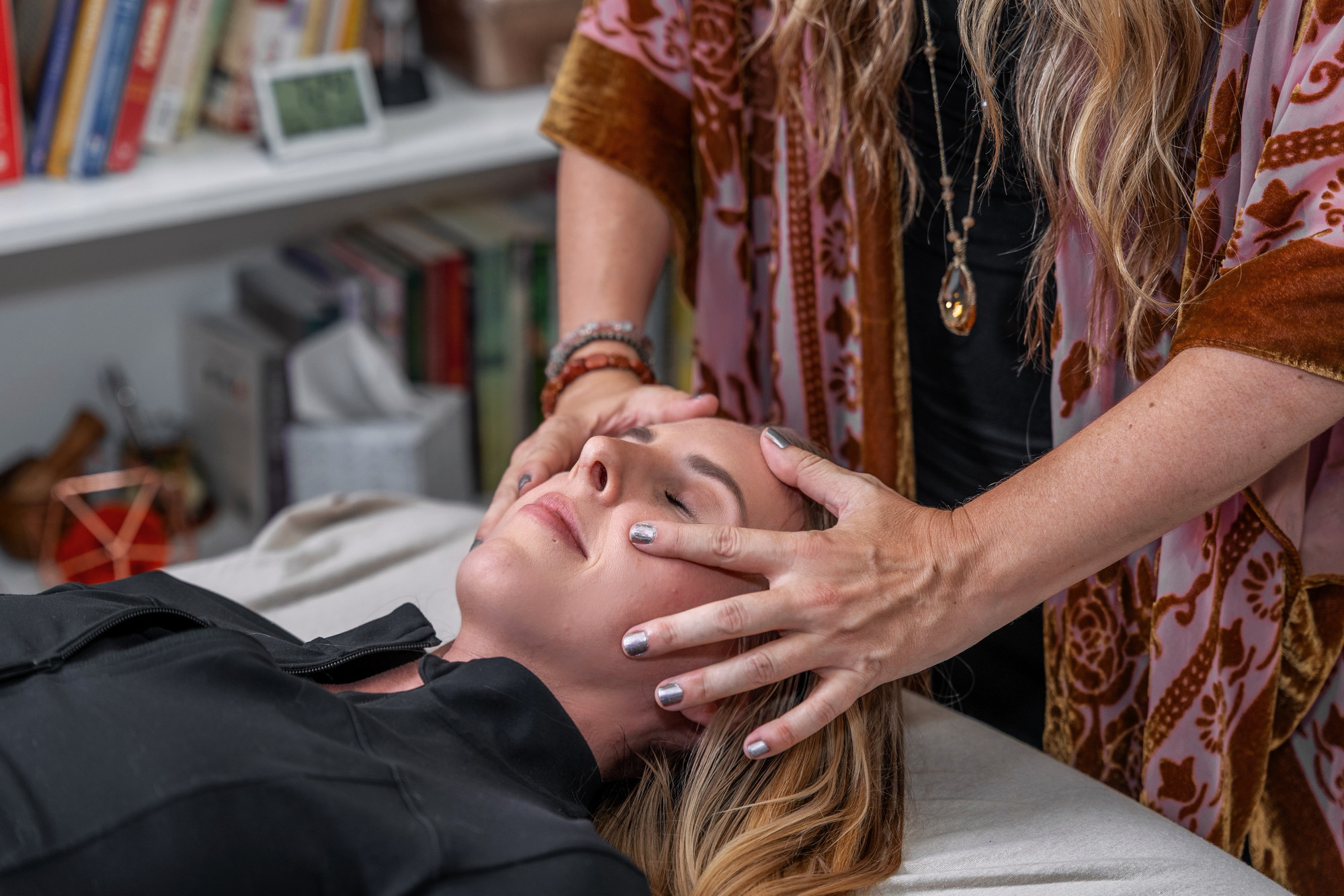 Person receiving a facial massage with eyes closed, lying on a treatment table, in a room with shelves and books.