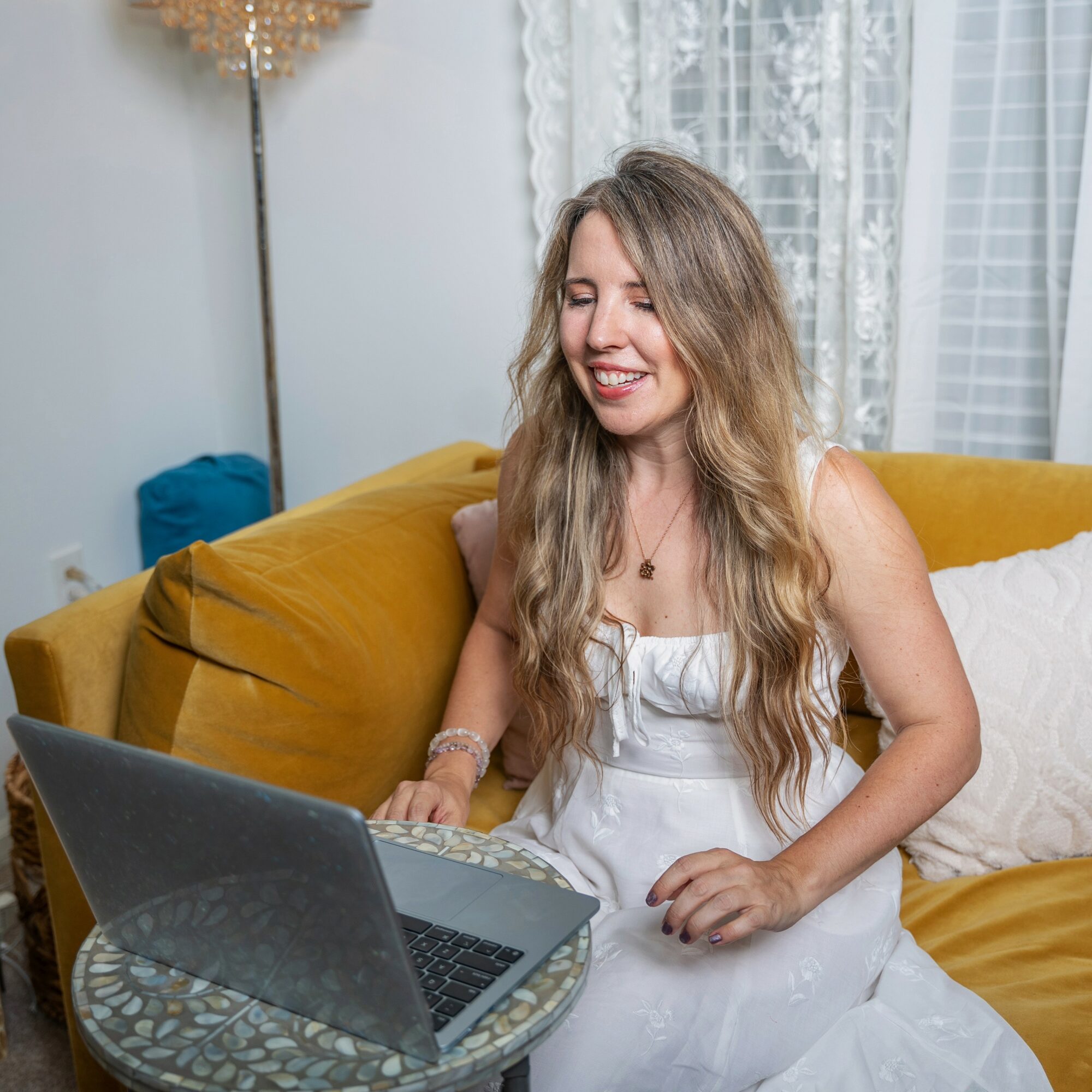Young woman with long wavy hair smiling while using a laptop on a yellow couch in a bright room.