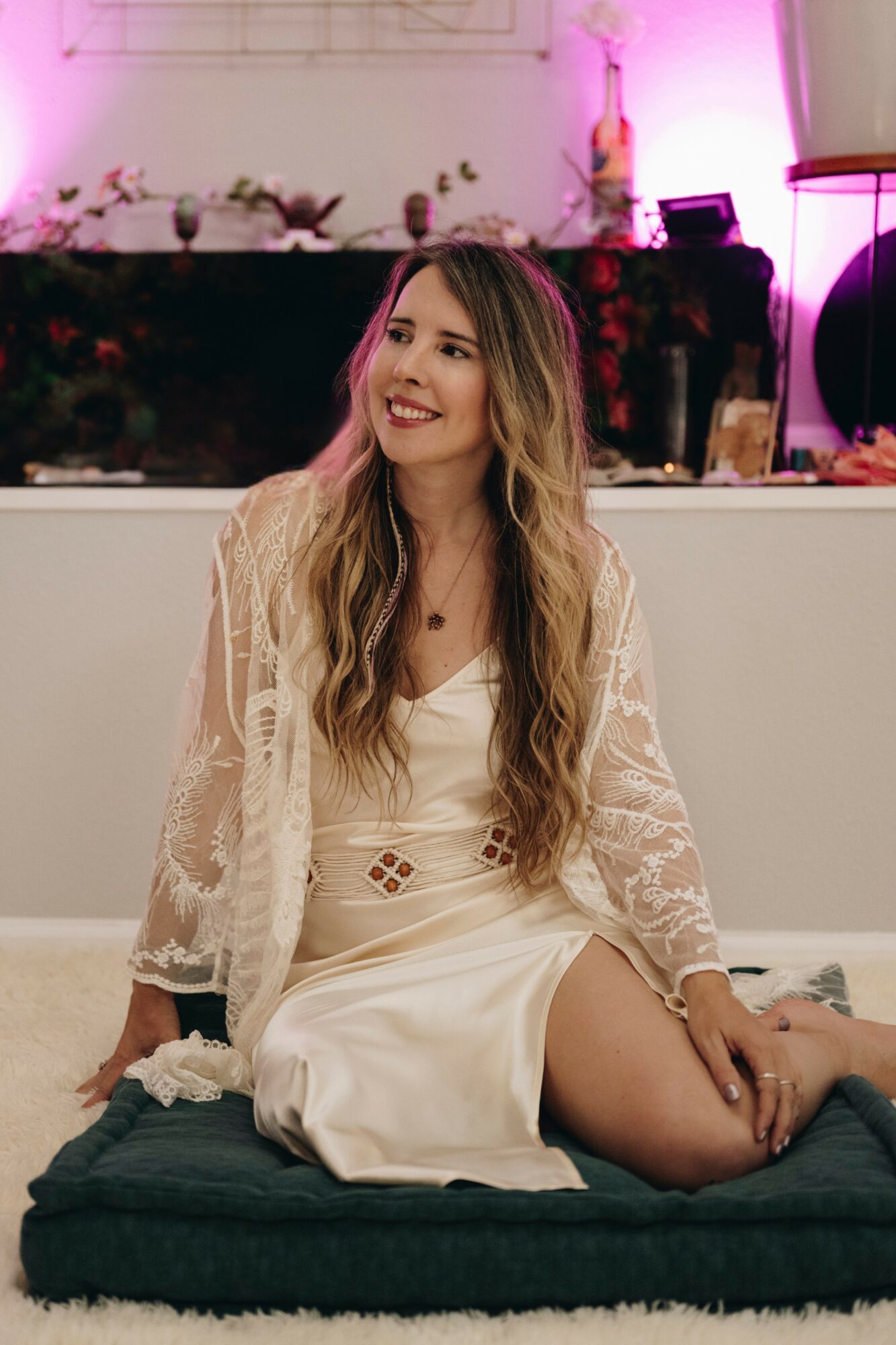 Woman with long hair smiling, sitting on a cushion in a decorated indoor space, wearing a white dress and lace cardigan.