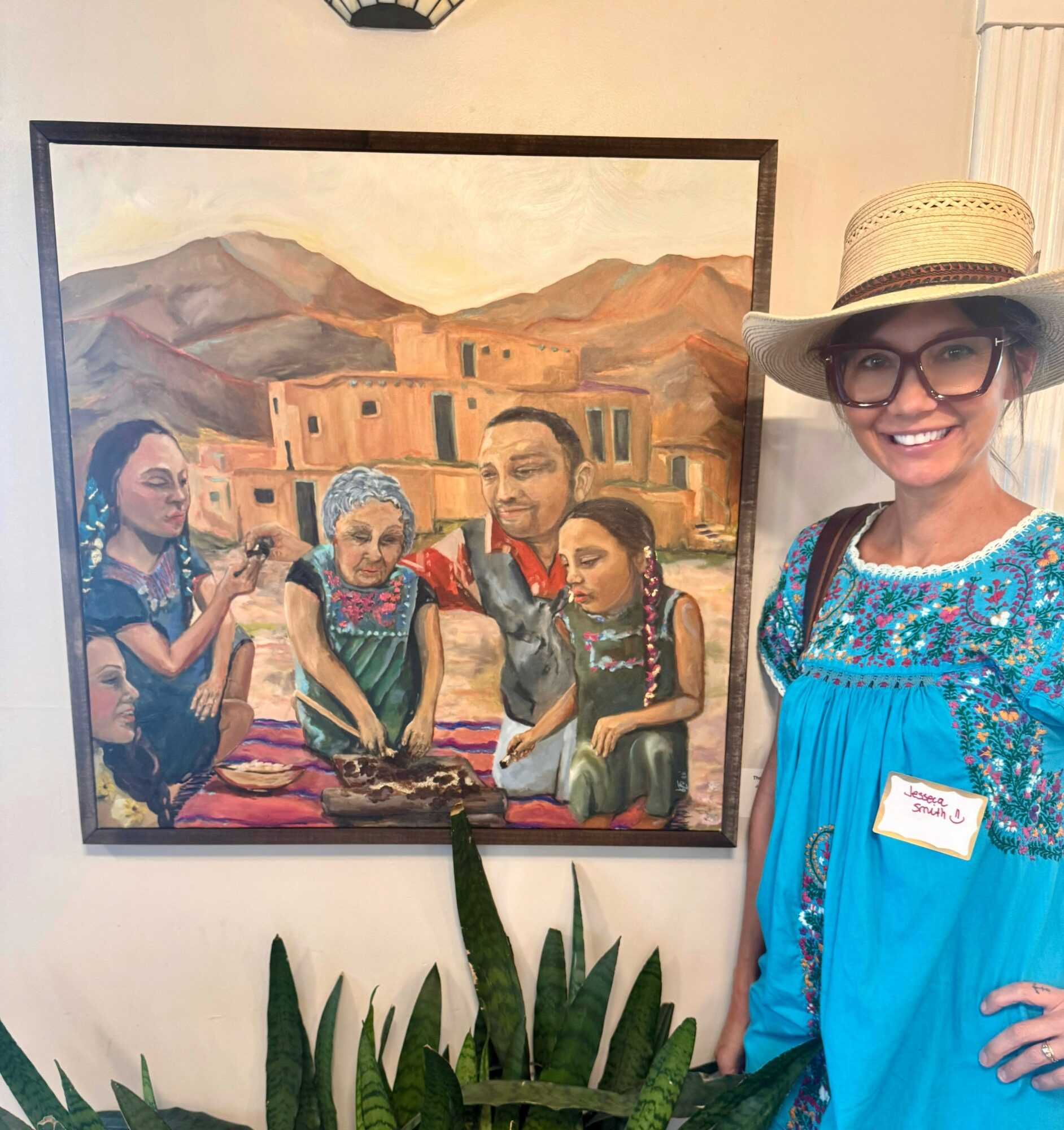 Woman in blue dress and straw hat smiling next to a painting of five people in a desert landscape.