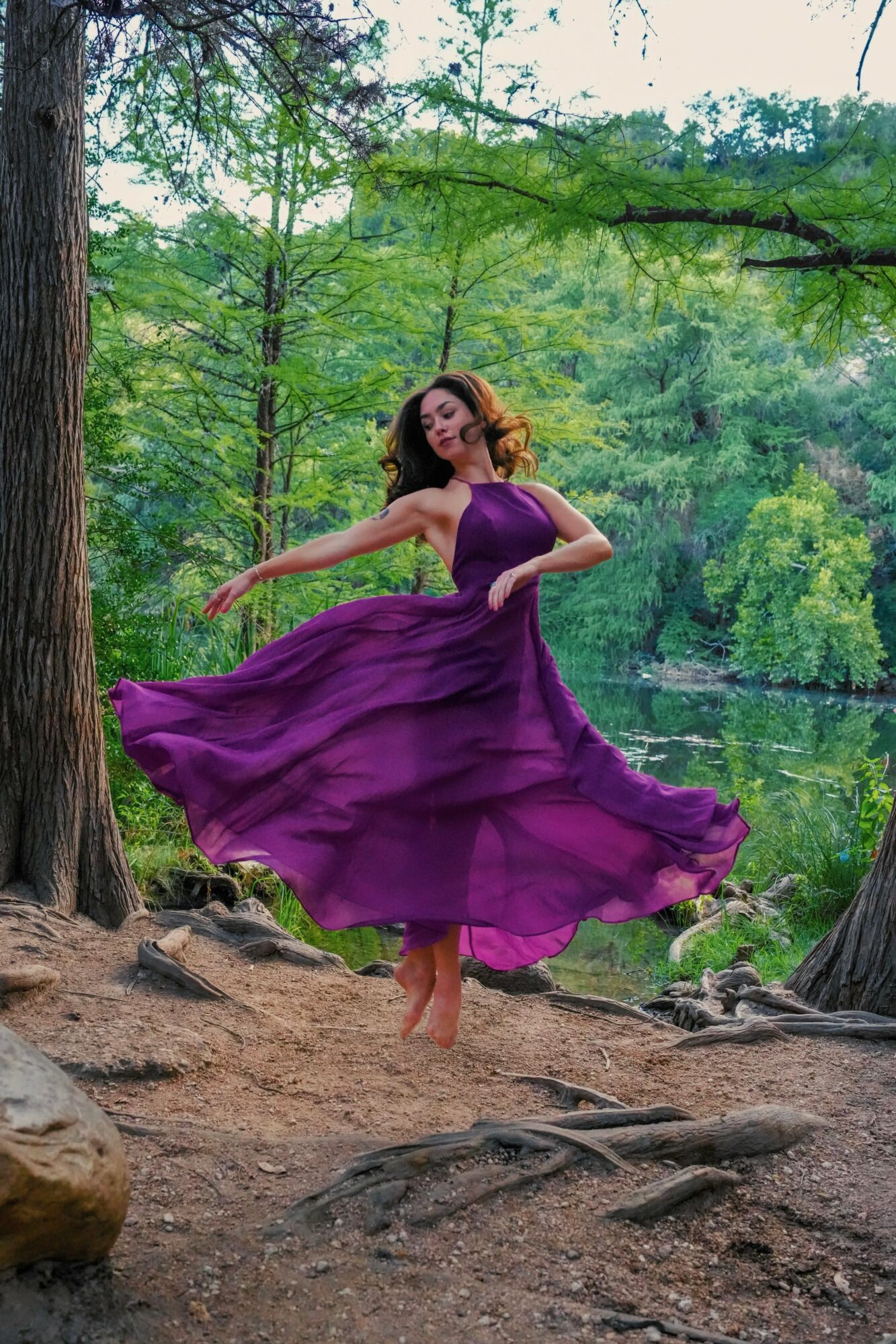 Woman in a flowing purple dress dancing outdoors near a lake surrounded by trees.