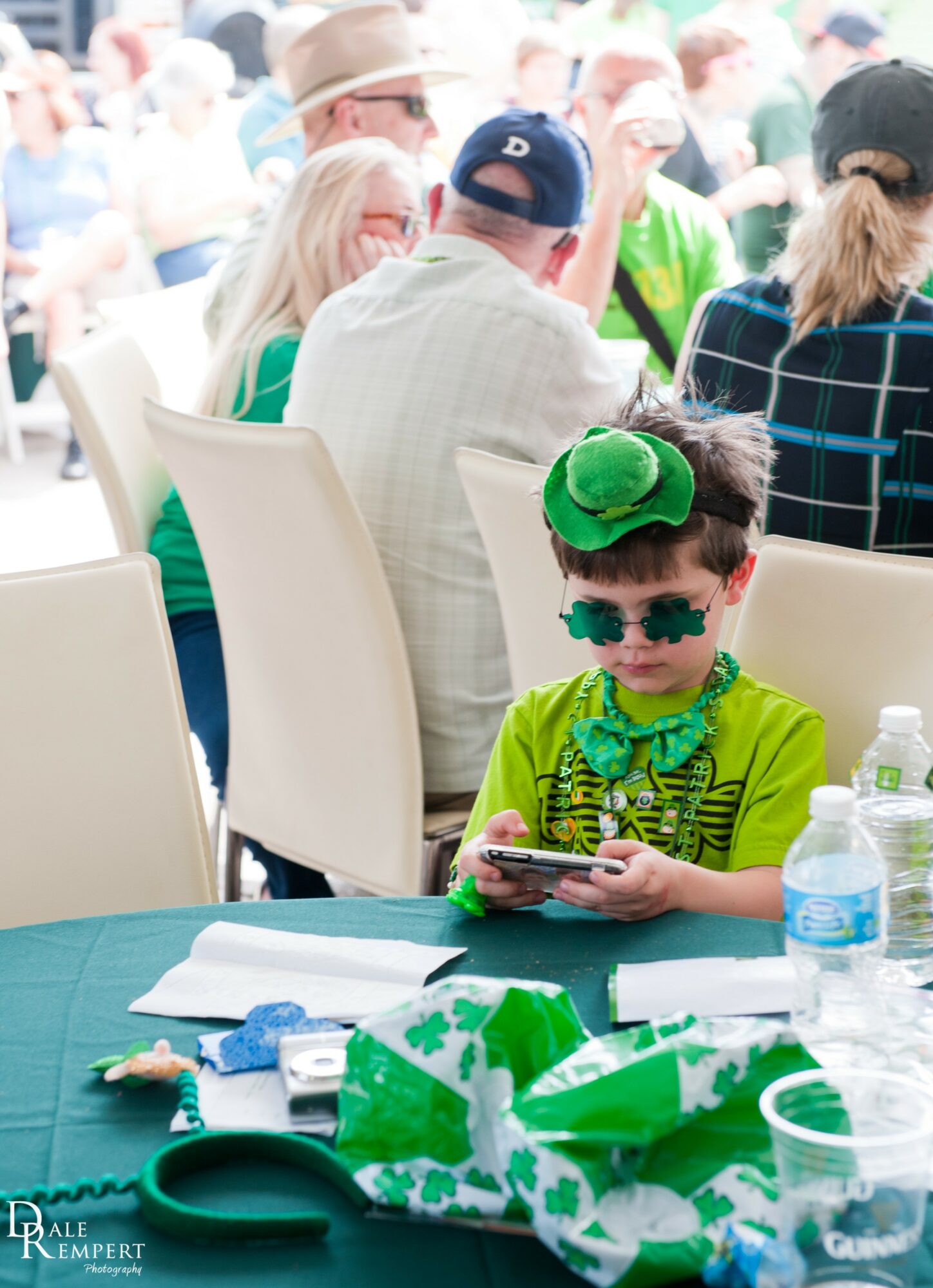 Child dressed as the Grinch wearing sunglasses and a green hat, sitting at a table with a phone, surrounded by water bottles.