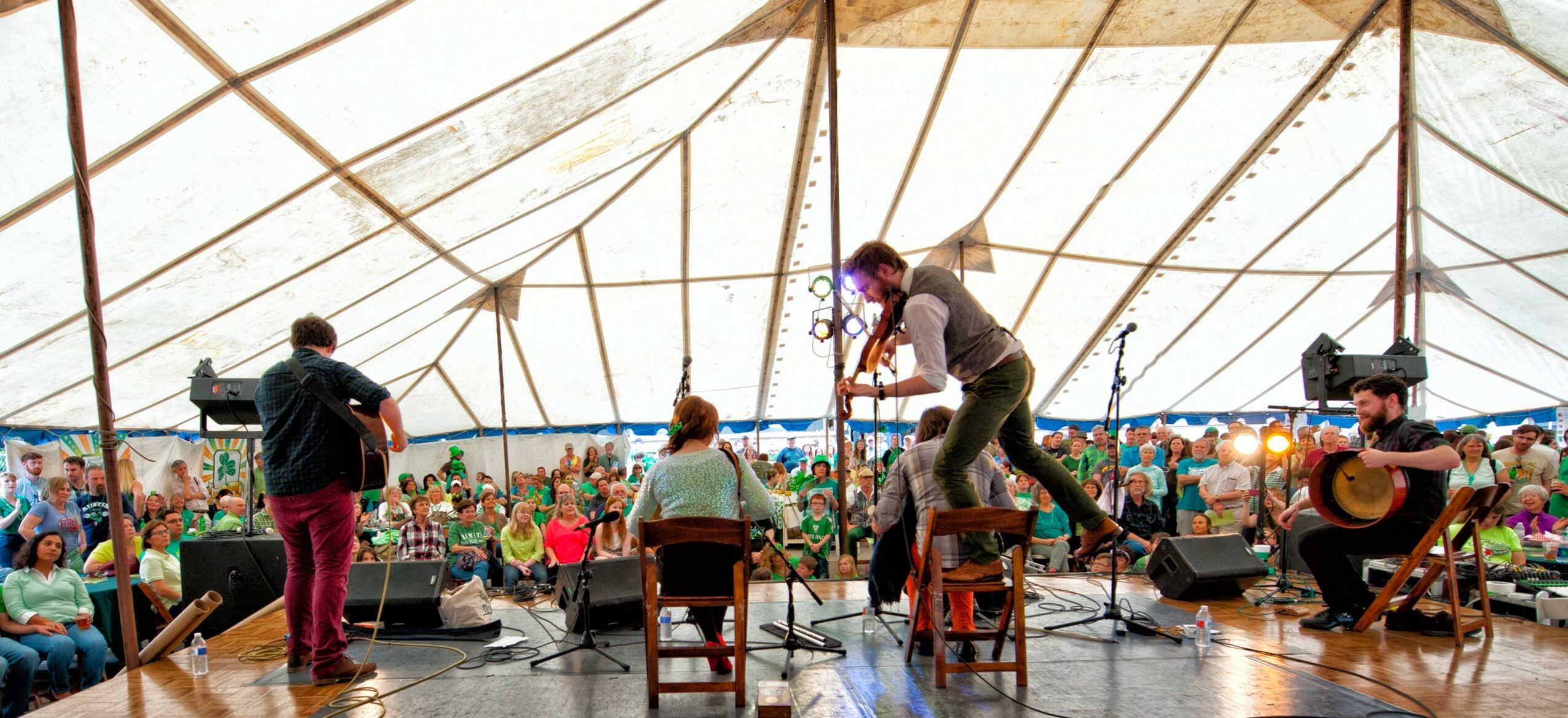 Musicians perform on stage inside a large tent with an audience watching outside, some seated and some standing.